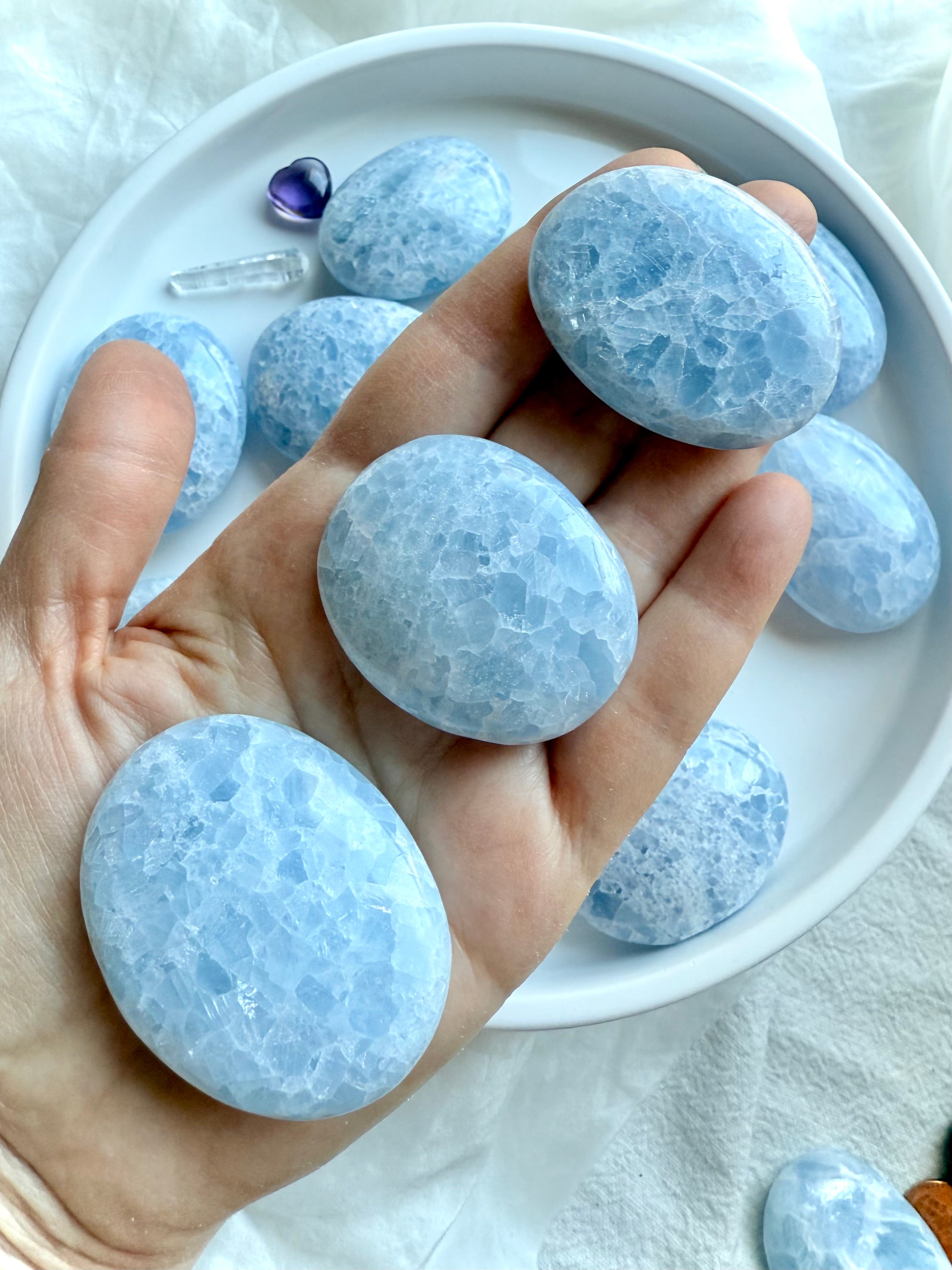 Three Blue Calcite palm stones shown on hand with more on a white plate below — soothing blue crystals for calm and clarity