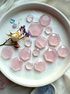 Flatlay of rose quartz hexagon crystals on a white plate styled with roses, clear quartz point and aquamarine moon, sacred geometry carvings for grids