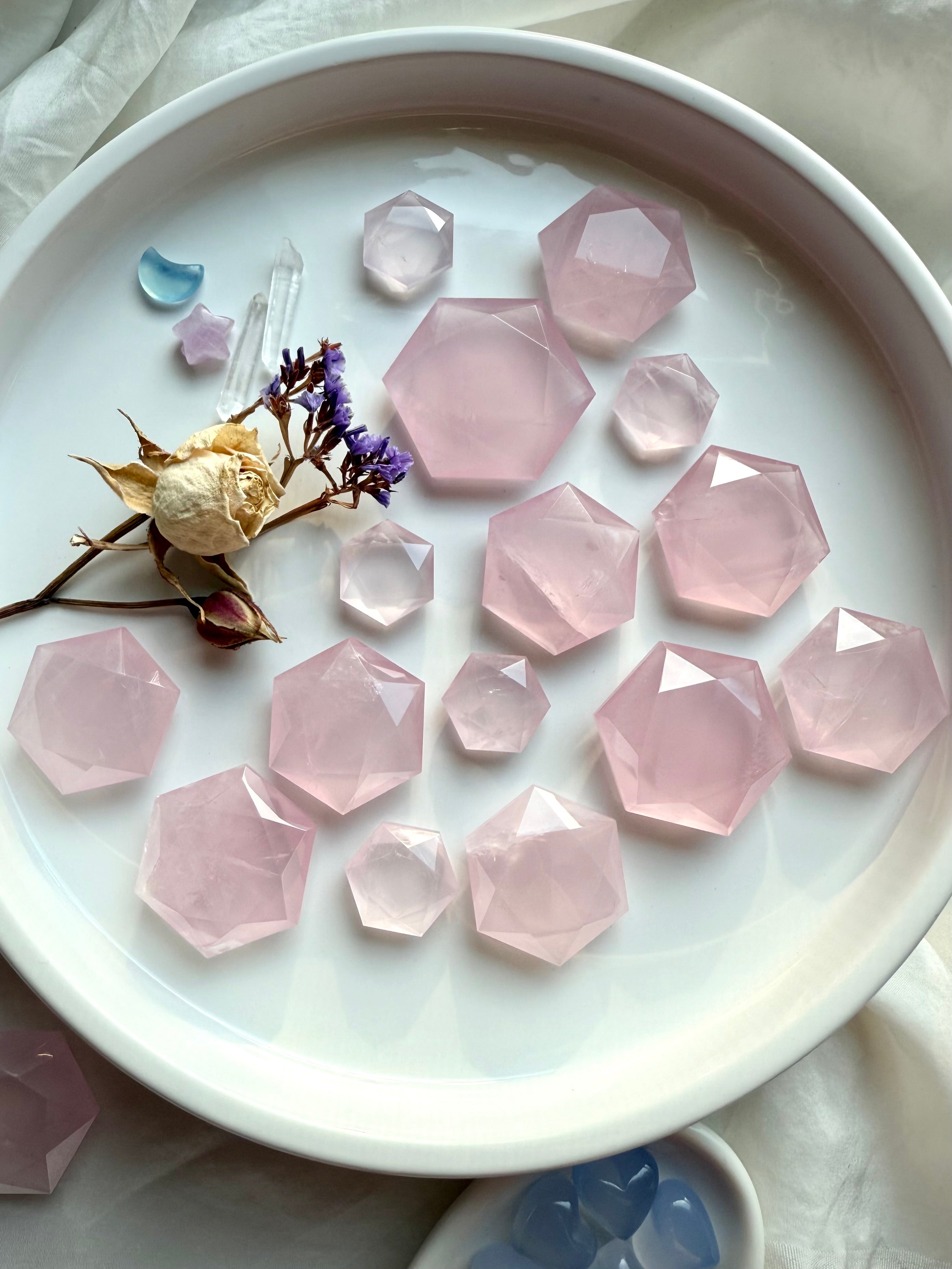 Flatlay of rose quartz hexagon crystals on a white plate styled with roses, clear quartz point and aquamarine moon, sacred geometry carvings for grids