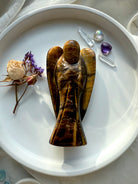 One-of-a-kind Tiger’s Eye angel carving with rich golden chatoyancy, displayed in a white dish with dried flowers and chakra crystals