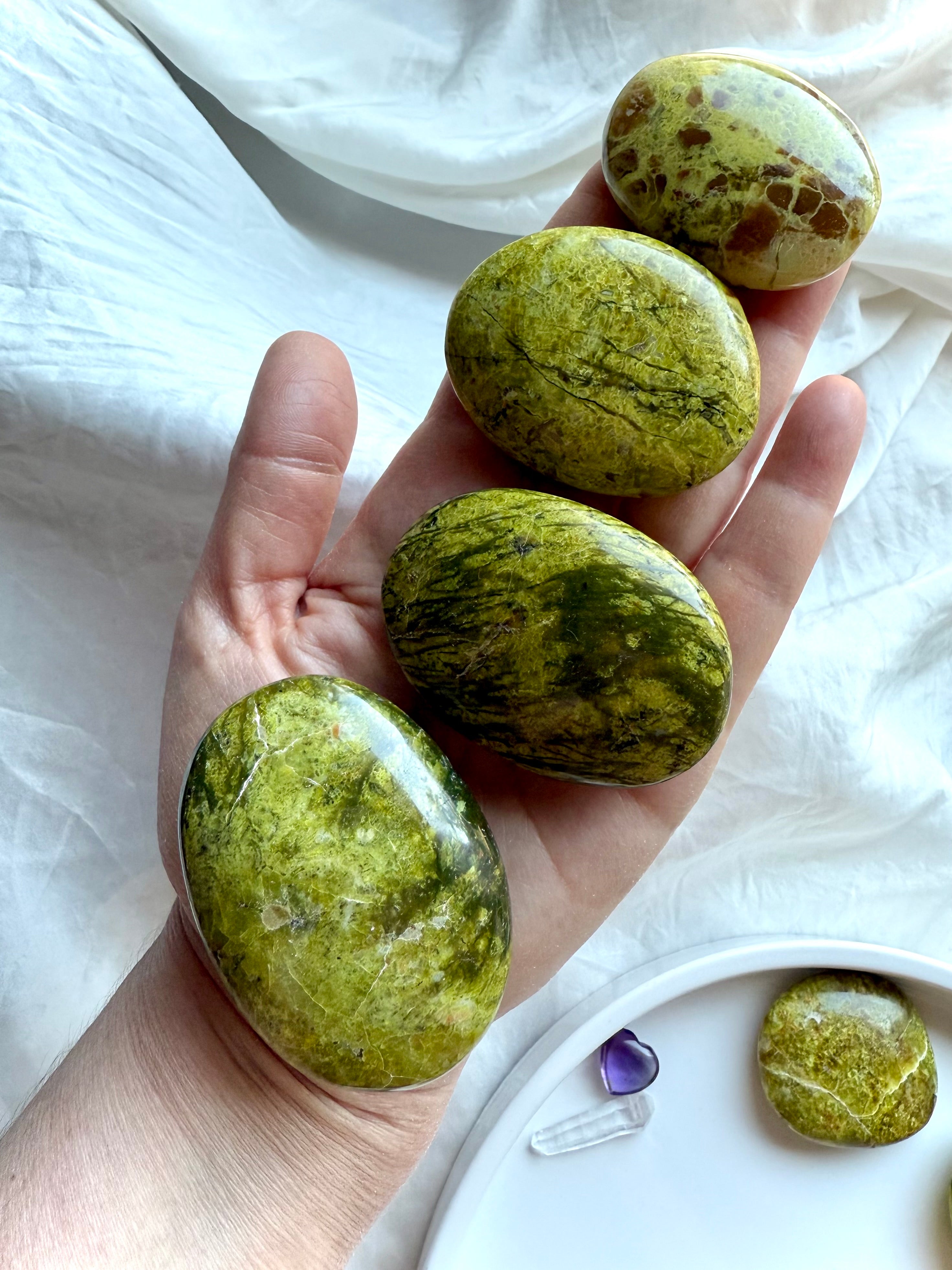 Four Green Opal palm stones displayed on a hand with stones below on white silk — heart chakra crystals for emotional balance and renewal.