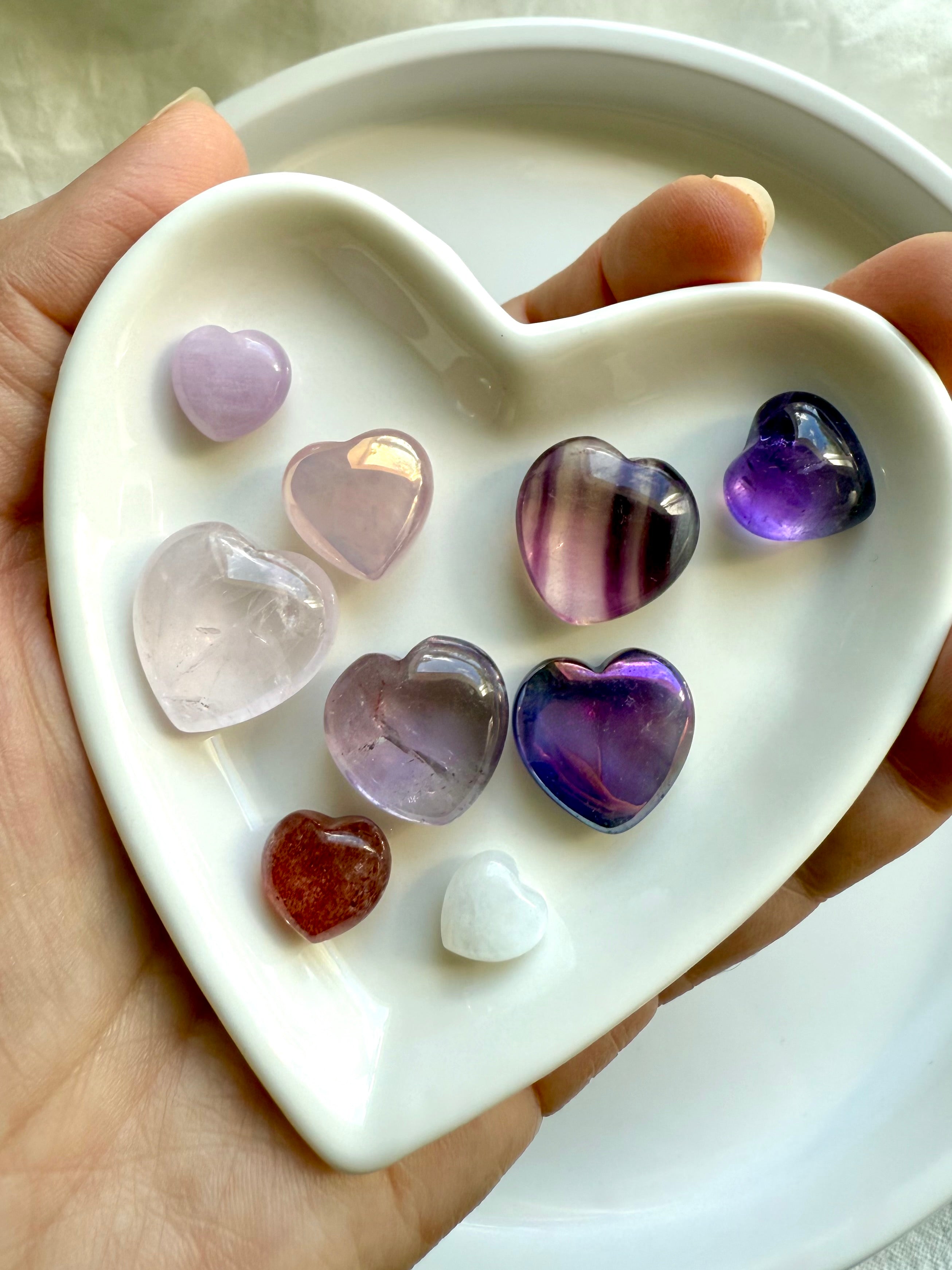 Close-up of white heart-shaped bowl holding nine pink and purple crystal hearts, premium mini gemstone carvings, held in hand.