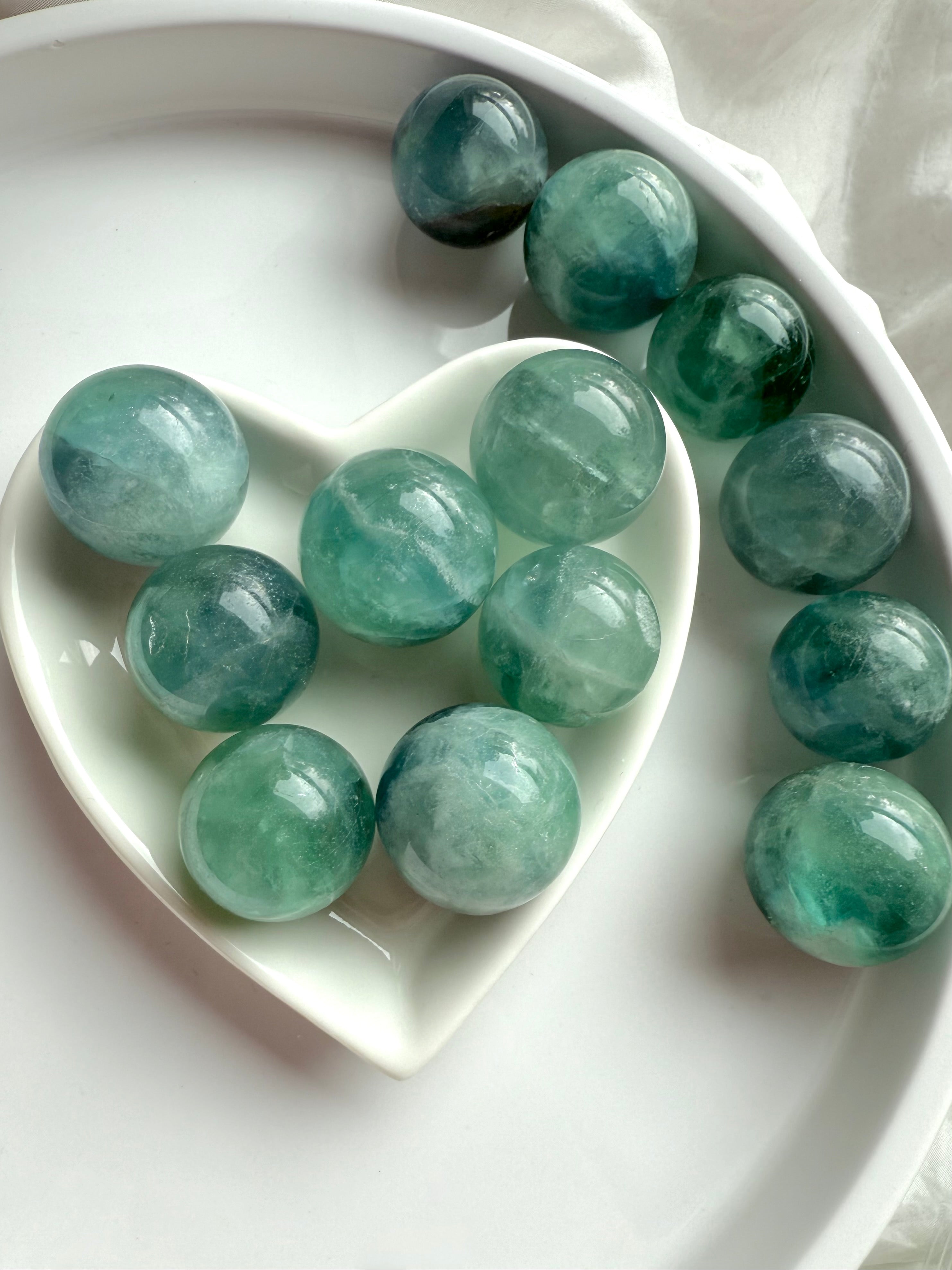Mini teal Fluorite spheres in a white heart dish, radiant aqua blue crystal balls for meditation and chakra energy work.