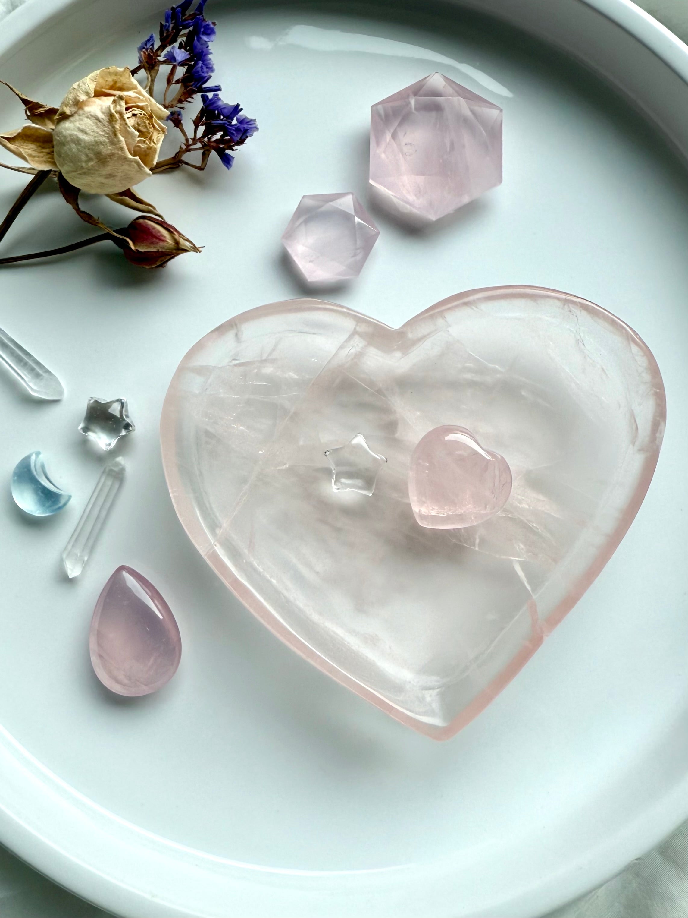 Rose Quartz heart-shaped crystal bowl displayed on a white tray with dried flowers and small gemstones, symbolizing love, healing, and heart chakra energy.
