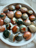 Mini Polychrome Jasper palm stones from Madagascar in a white bowl, showcasing earthy red, green, cream, gray, and brown landscape patterns