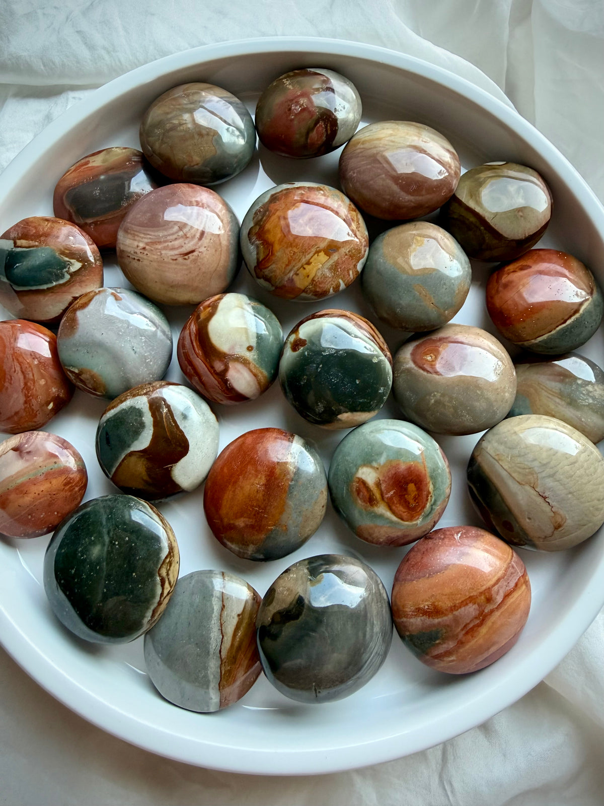Mini Polychrome Jasper palm stones from Madagascar in a white bowl, showcasing earthy red, green, cream, gray, and brown landscape patterns