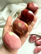 hand holding smooth Mookaite Jasper palm stones with natural pink, cream, and deep red banding for grounding energy