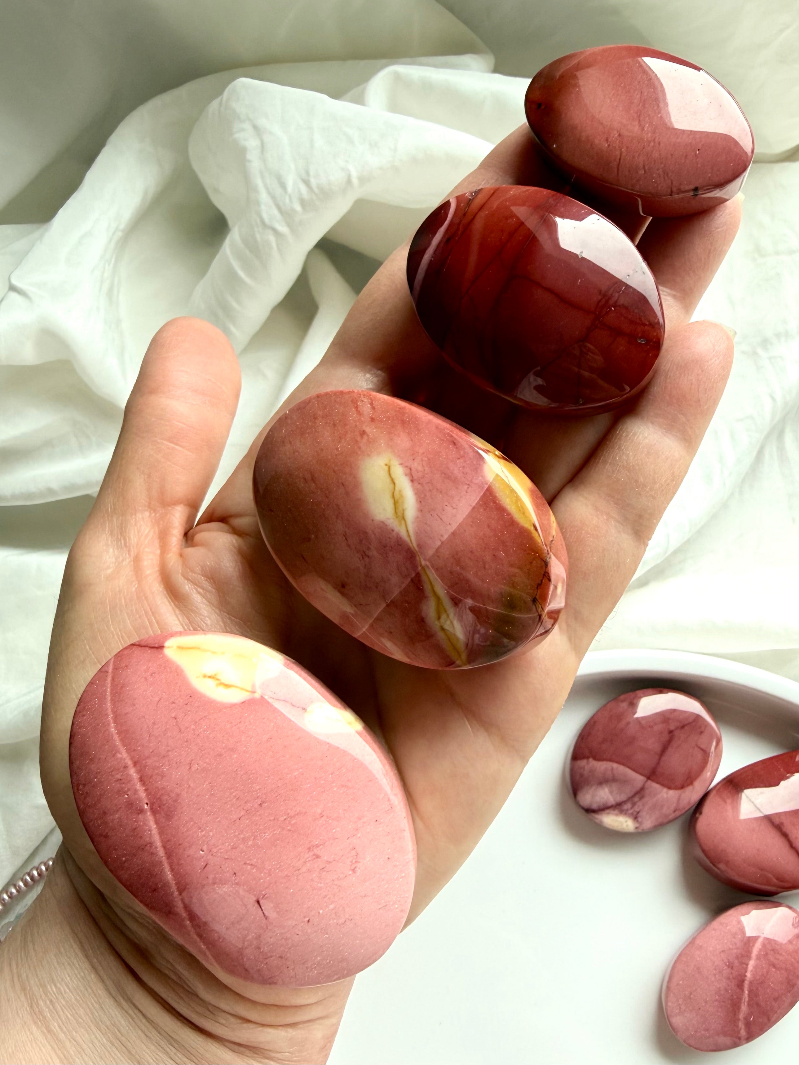hand holding smooth Mookaite Jasper palm stones with natural pink, cream, and deep red banding for grounding energy
