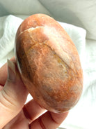 Hand holding a polished Peach Moonstone palm stone with soft natural shimmer and warm earthy peach coloring