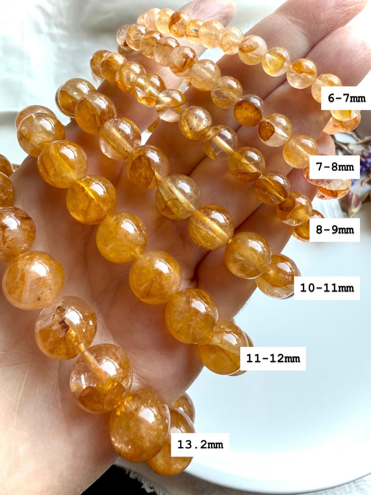Golden Healer Quartz bracelet stack on hand with bead sizes labeled 6–7mm through 13.2mm, showing variation in size and glowing golden hues.
