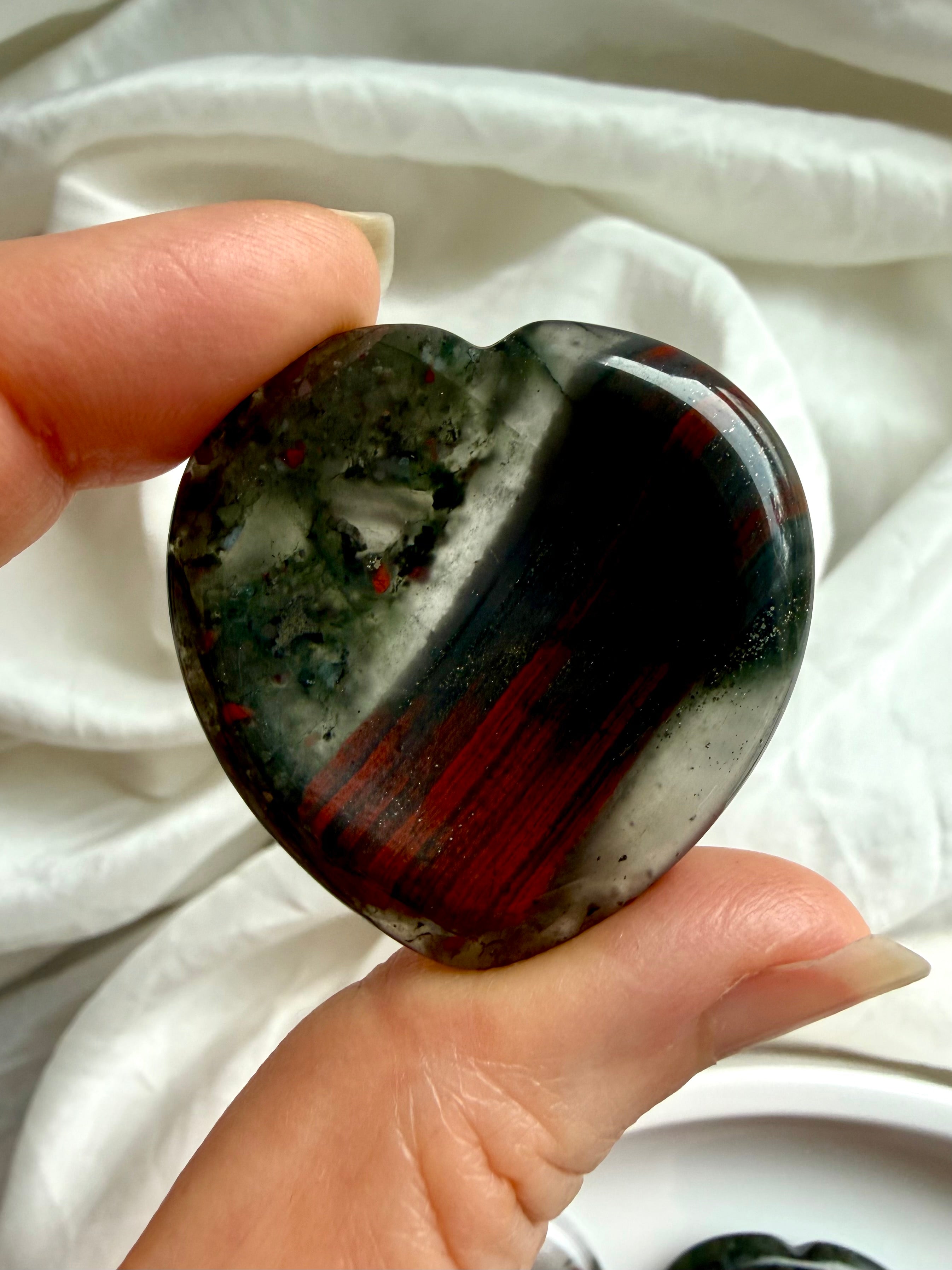 close-up bloodstone heart worry stone held between fingers showing natural pattern variation, crystal for courage stability and stress relief