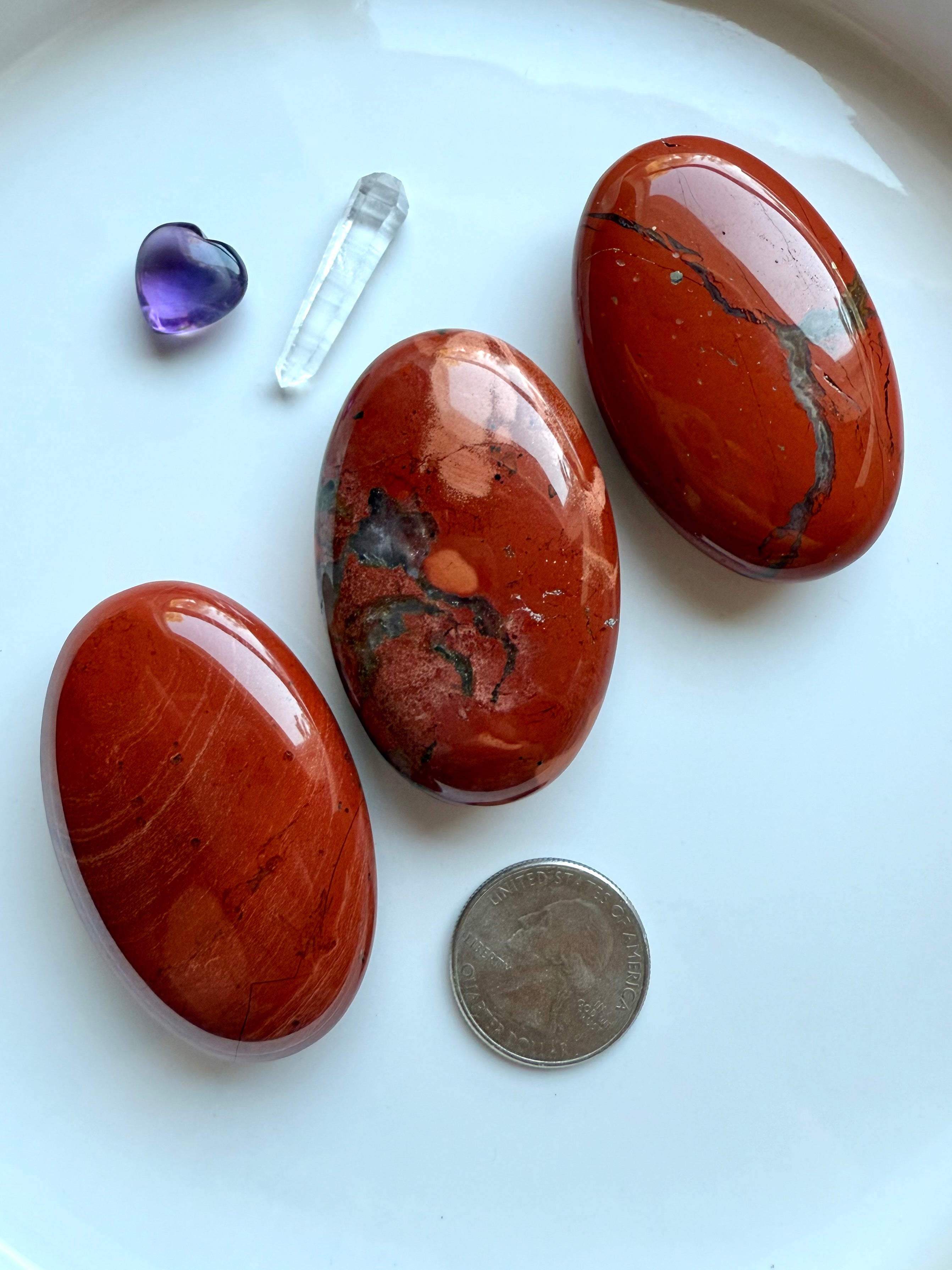 Three Red Jasper palm stones on a white plate with quarter for scale and amethyst heart carving.