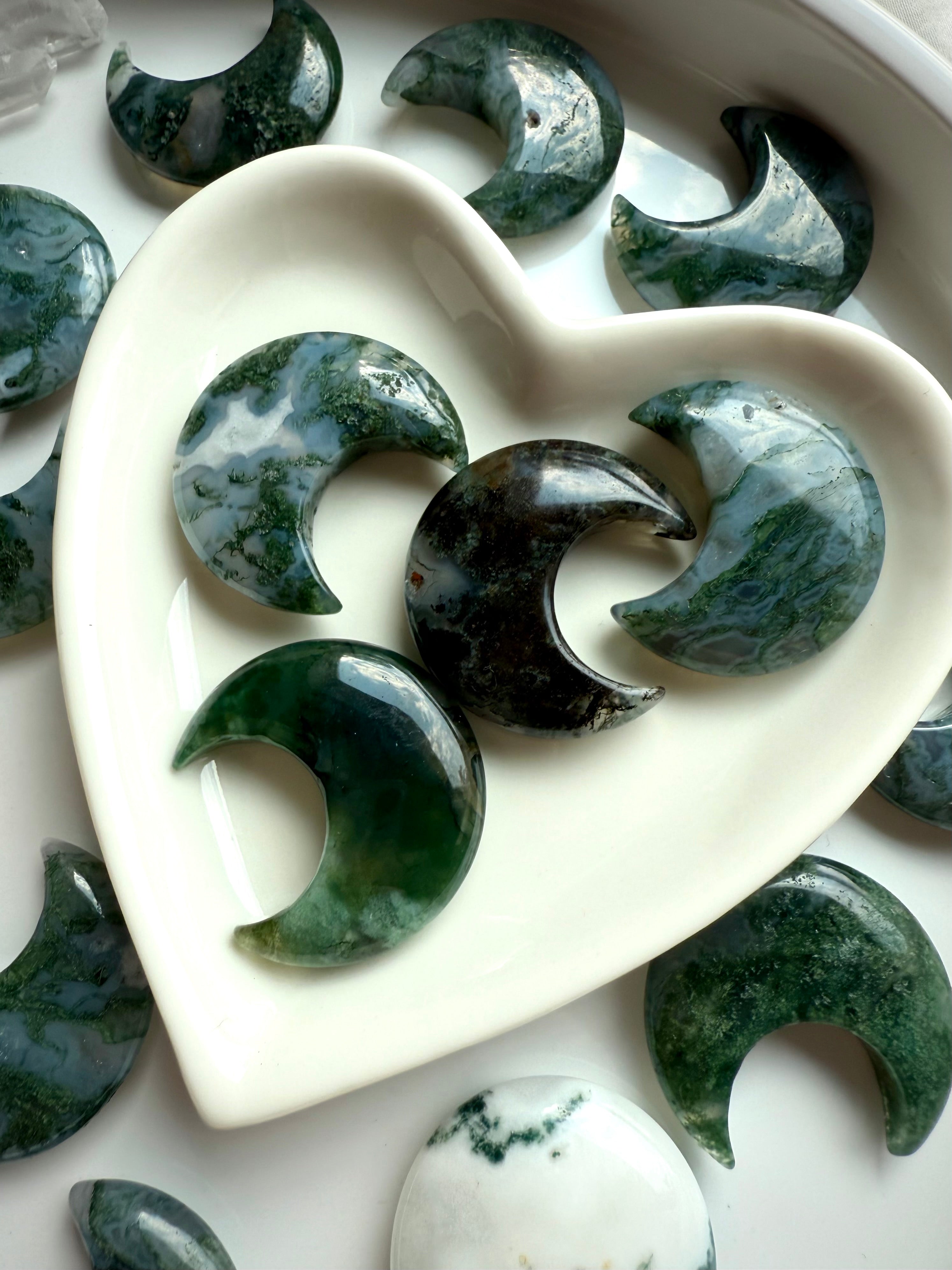 Detailed view of Moss Agate moon carvings in a white heart dish, highlighting natural mossy inclusions and earthy green tones.