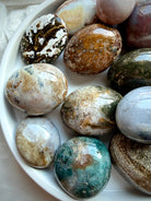 Ocean Jasper palm stones in a white bowl showing a diverse assortment of earthy colors, natural banding, and orbicular formations
