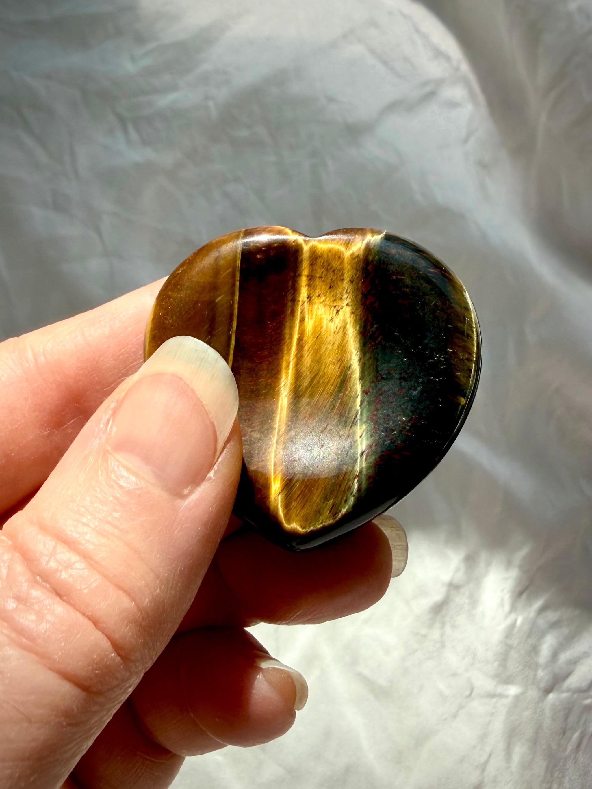 Tiger’s Eye heart worry stone held between fingers, revealing silky golden chatoyancy and deep brown mineral bands