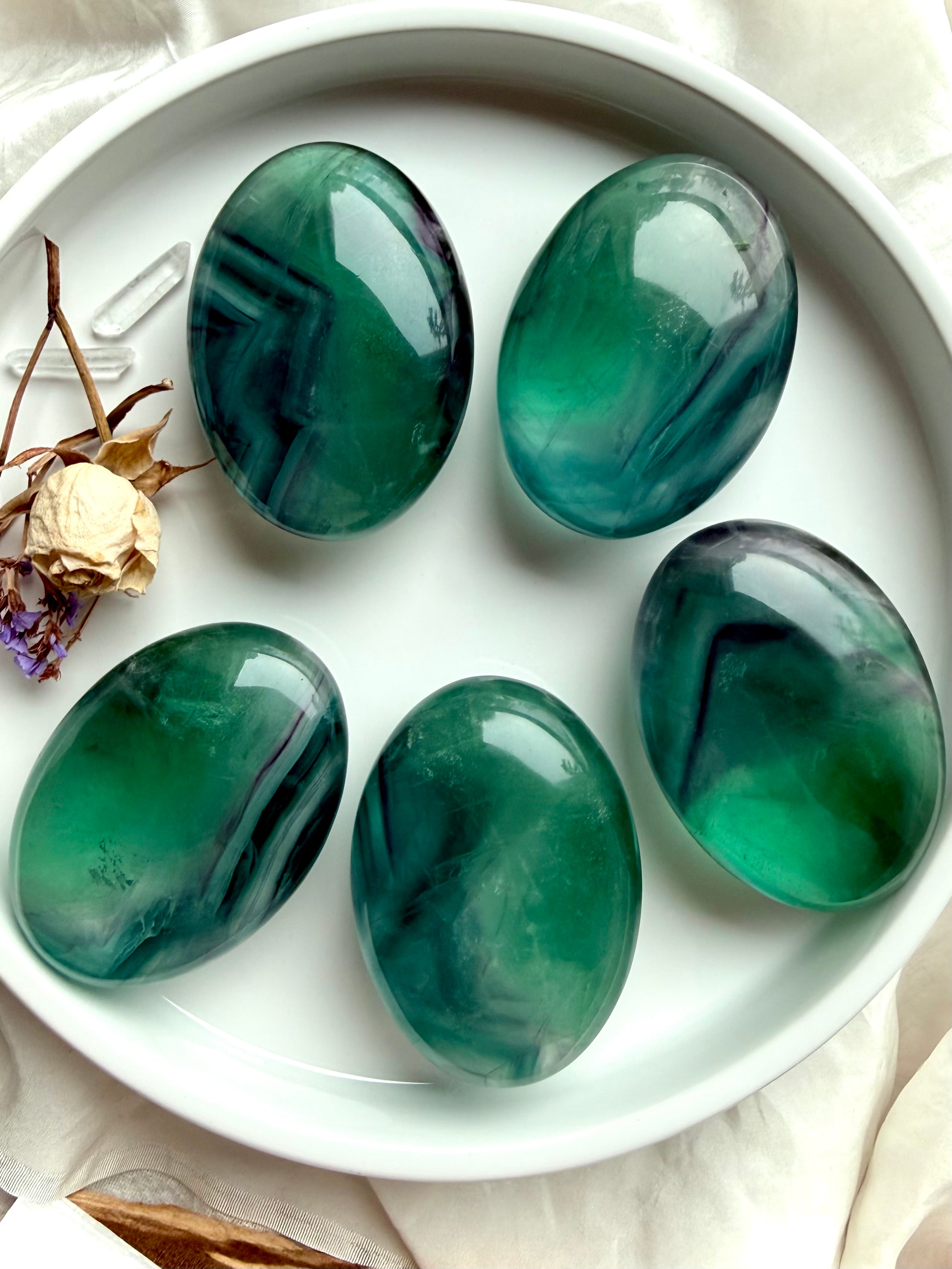 Large teal fluorite palm stones displayed on a white tray with dried florals, highlighting luminous blue green layers