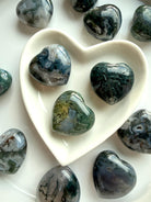Moss Agate crystal hearts in a white heart-shaped bowl, showing natural green moss inclusions and dreamy chalcedony patterns
