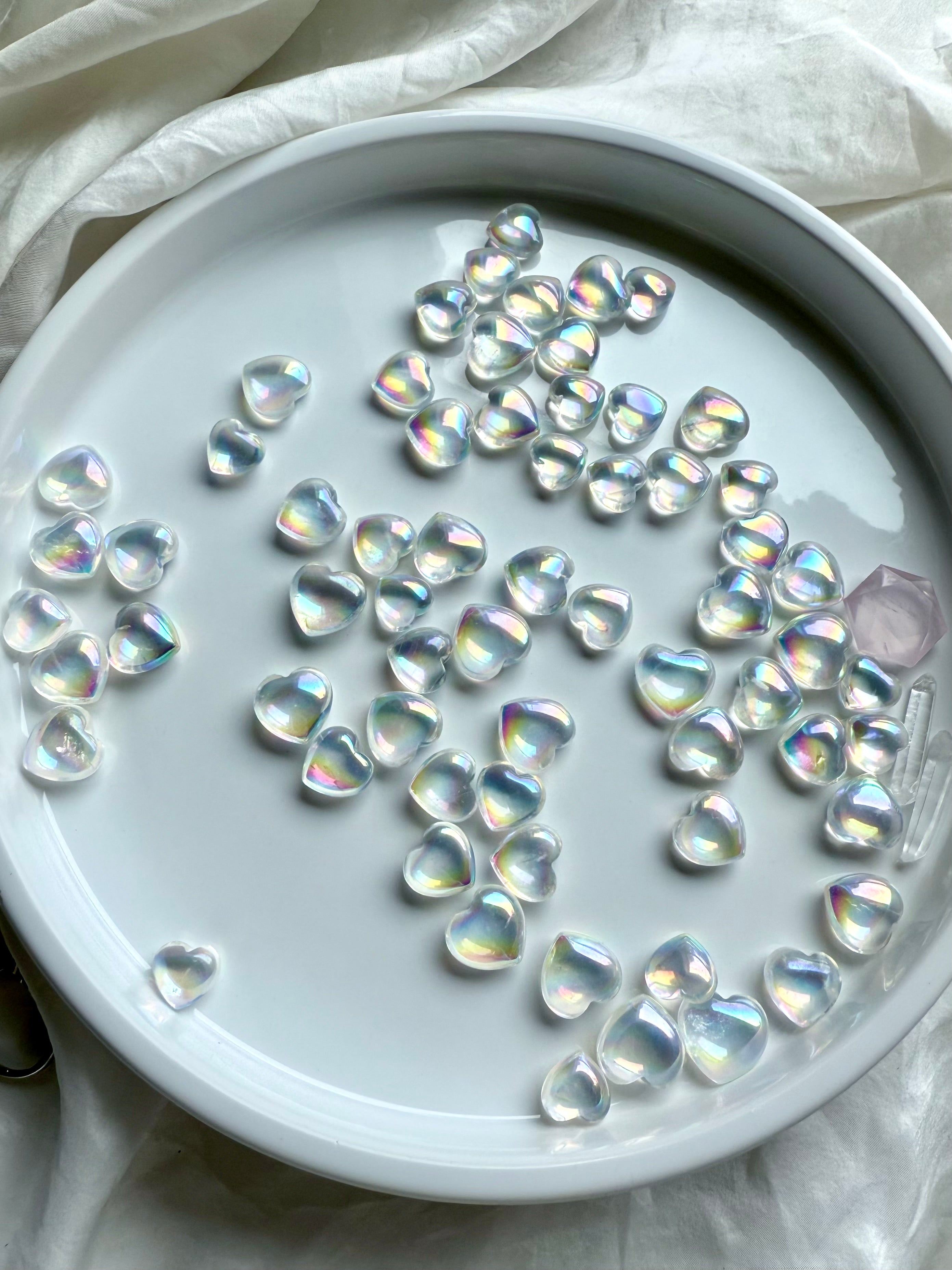 Dozens of Angel Aura Quartz mini hearts on a white plate with soft natural light, showing their rainbow sheen