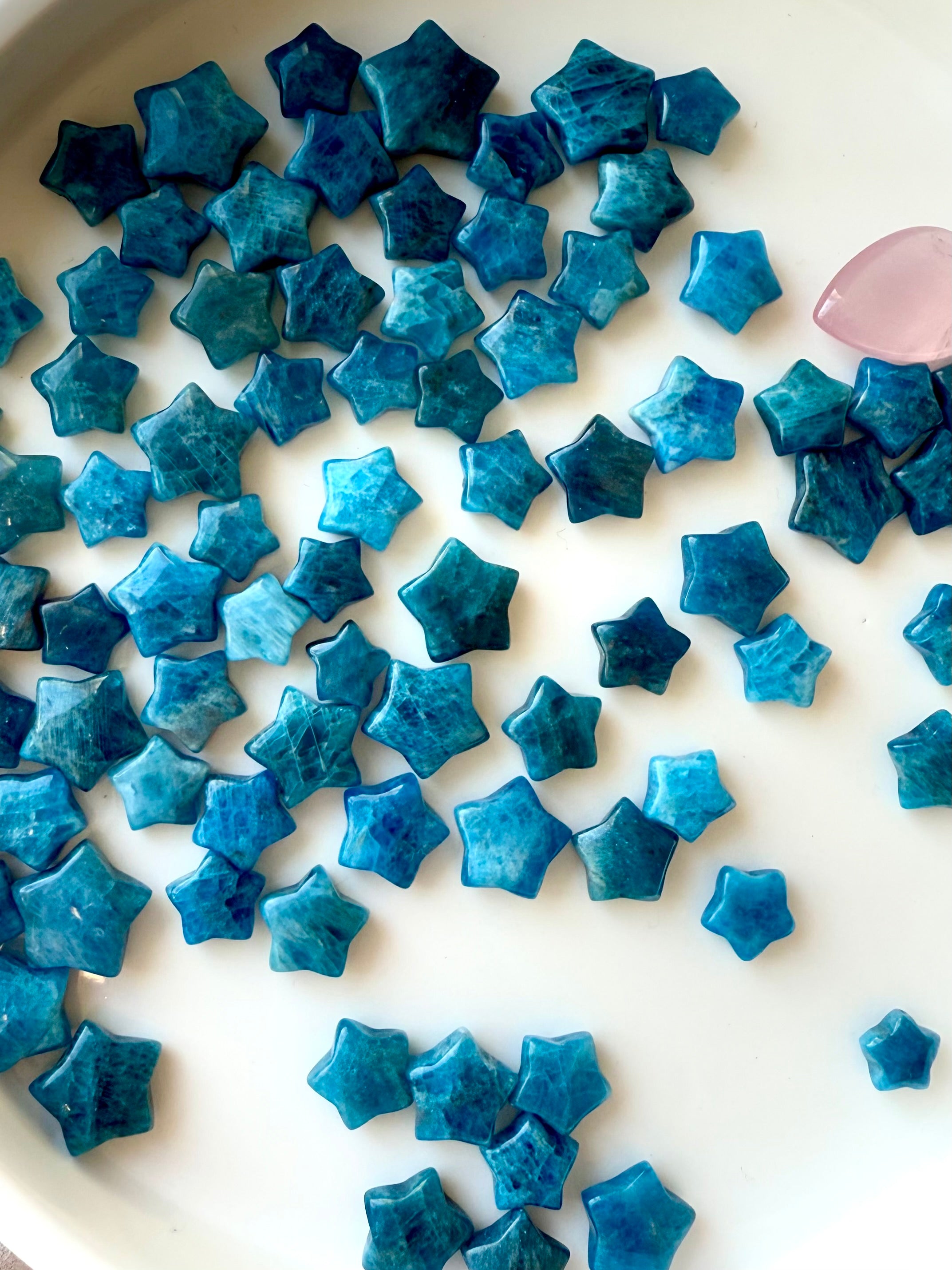 Overhead view of Blue Apatite crystal stars on white plate — mini teal star carvings showing size, hue, and texture variations.