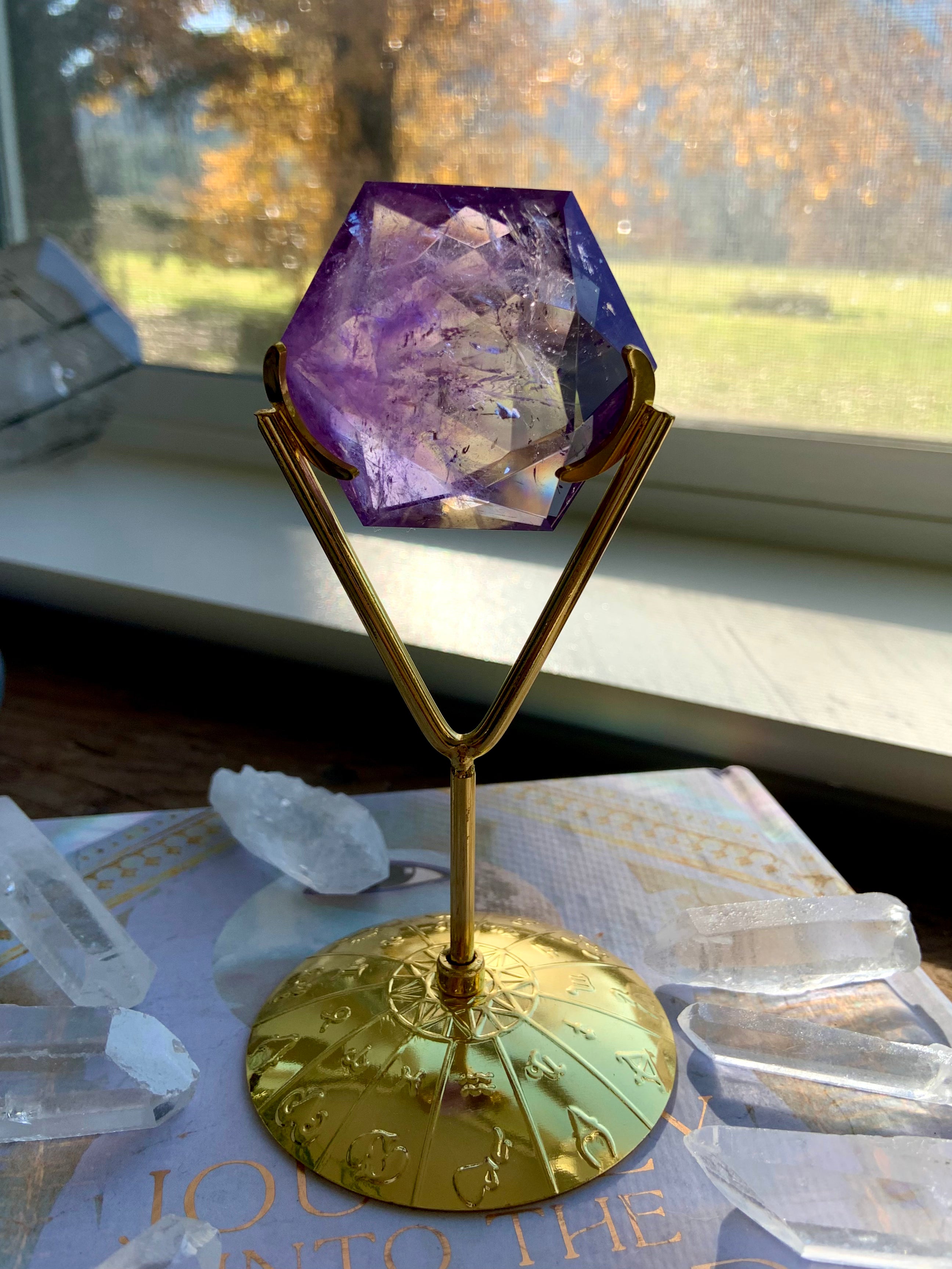 Lifestyle image of gold crystal stand holding Amethyst hexagon — window light illuminates the purple crystal with greenery in the background.