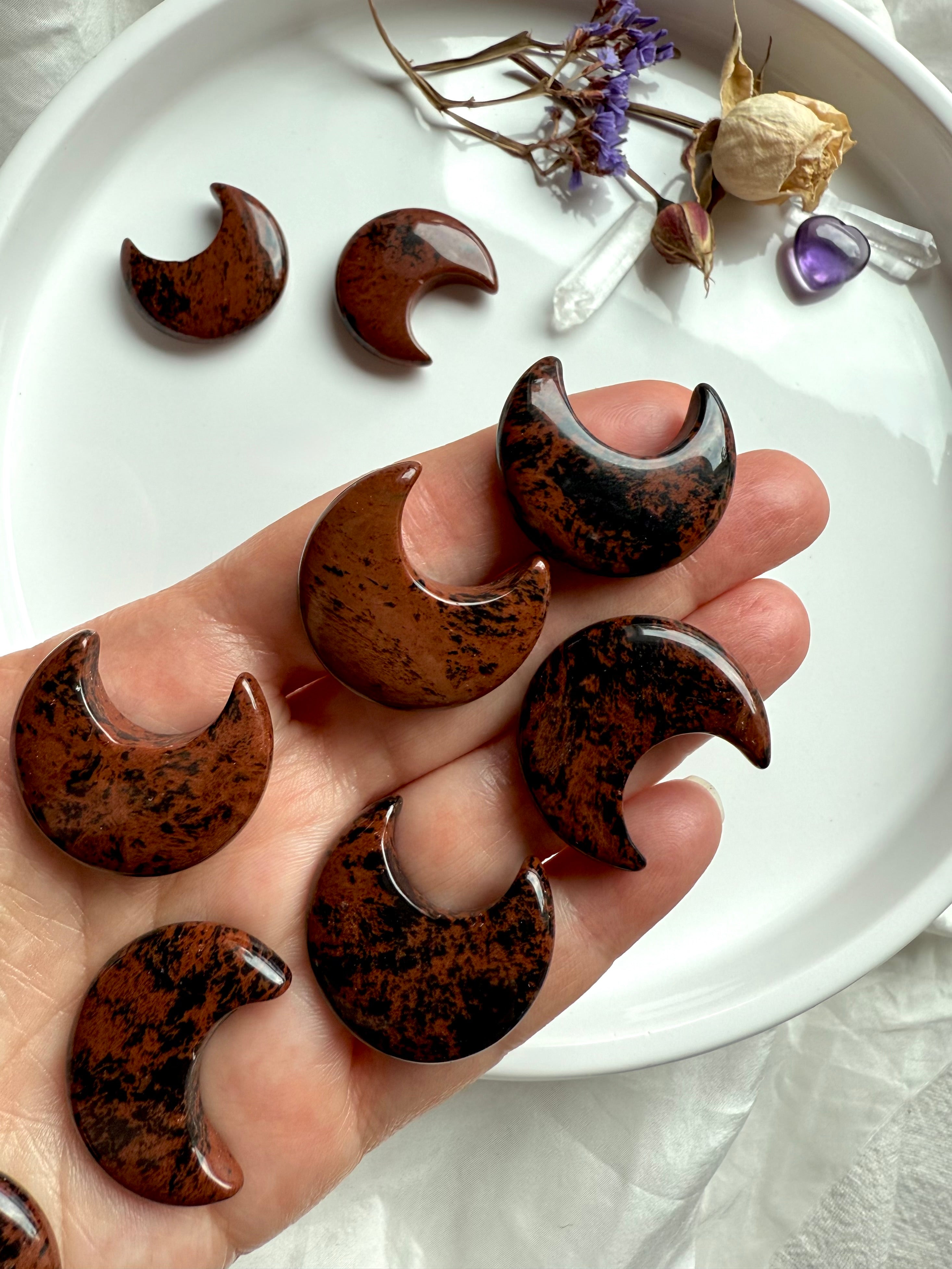 Mahogany Obsidian crescent moons held in hand, showing rich red-brown and black swirls. Grounding crystal for emotional strength.