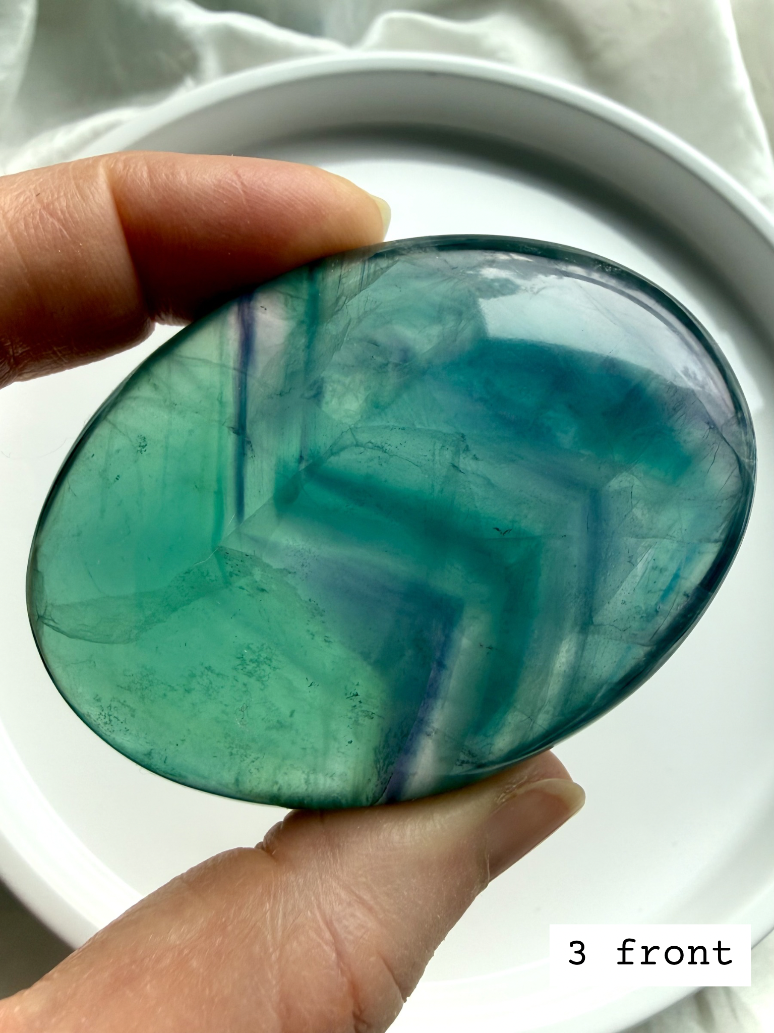 Teal fluorite palm stone #3 front held to the light, highlighting vivid blue green striping and clarity