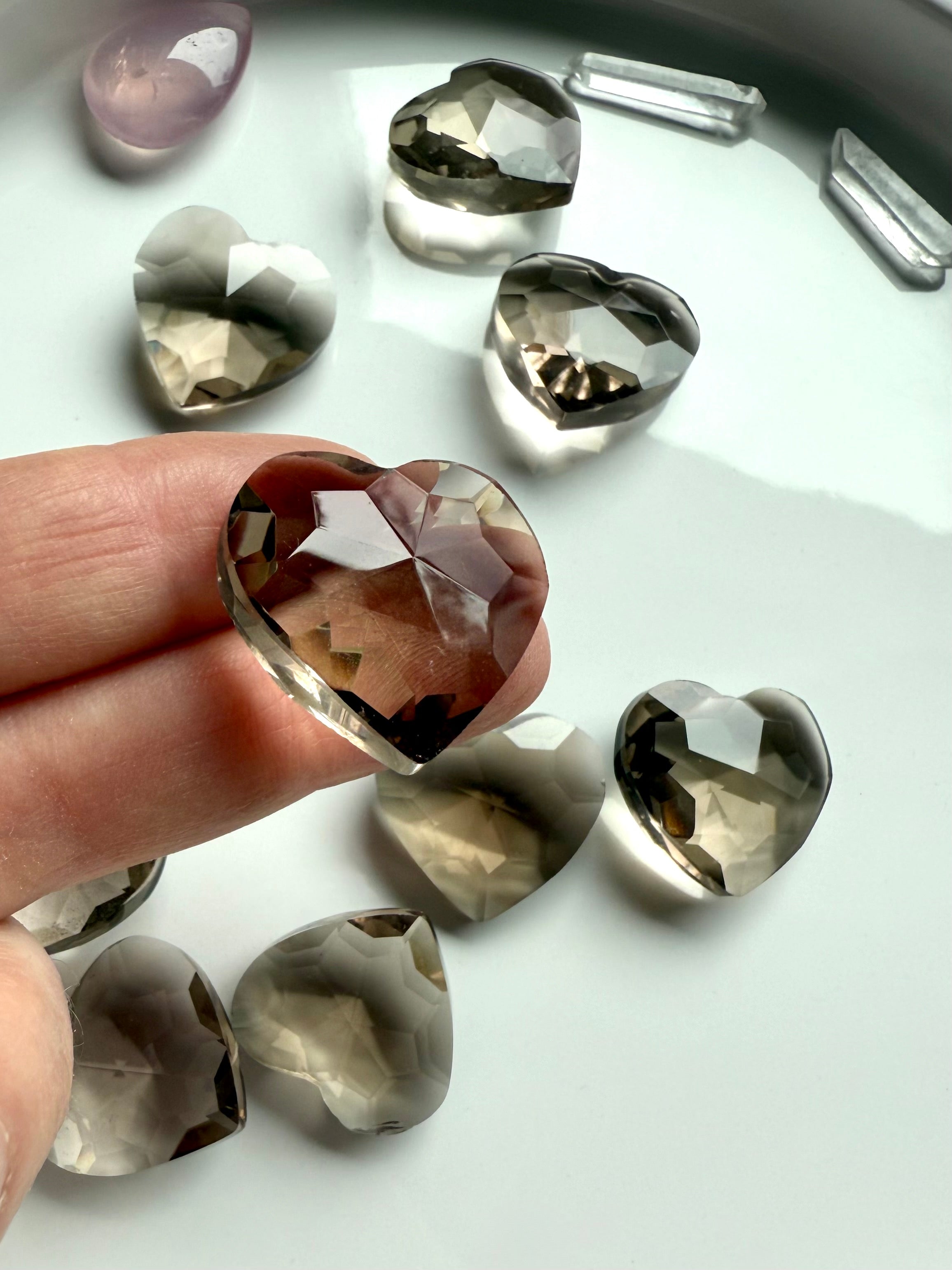 Faceted Smoky Quartz crystal heart held on fingertips with more hearts in soft focus — grounding crystal for energy clearing, emotional support, and healing.