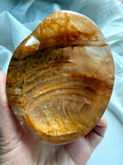 Hand-carved Golden Healer gemstone bowl held up to the light, revealing iron oxide bands, translucent girasol texture, and curved oval shape