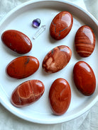 Flatlay of polished Red Jasper palm stones on a white plate — grounding Root Chakra crystals showing natural color and pattern variation.