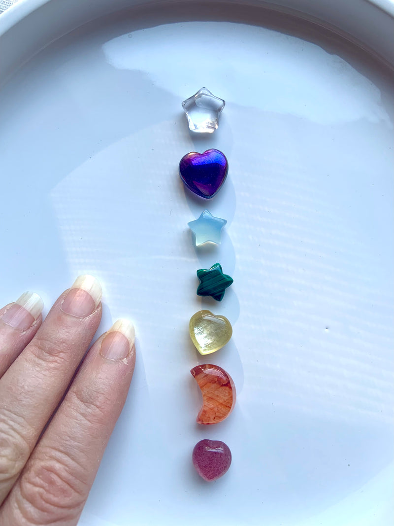 7 chakra crystal set of mini rainbow crystal moons stars and hearts shown on a white plate with my hand for scale