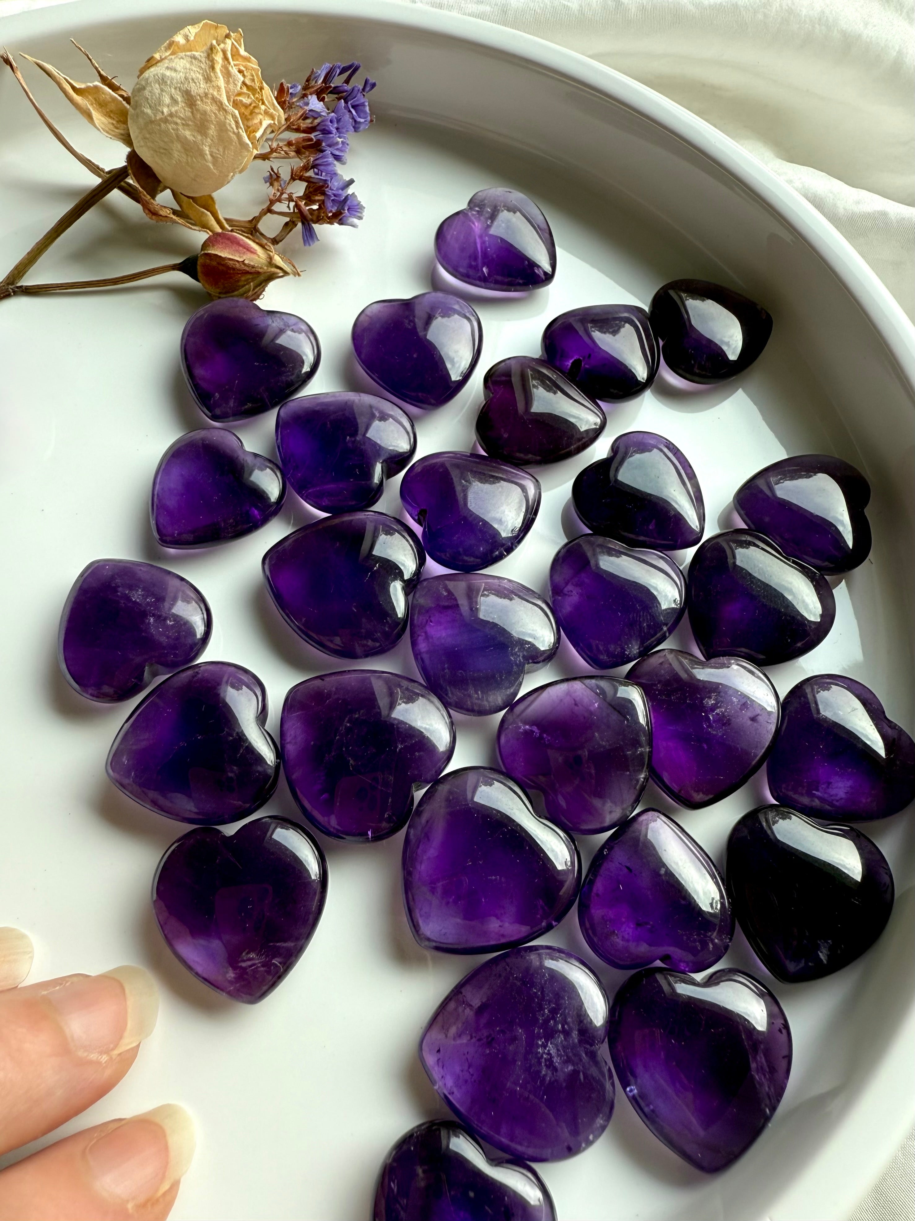 Amethyst crystal hearts in deep purple tones displayed in a dish with dried flowers — polished crown chakra stones for intuition, clarity, and divine guidance