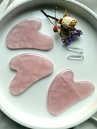 Three Rose Quartz crystal gua sha facial massage tools on white plate with flowers and quartz points