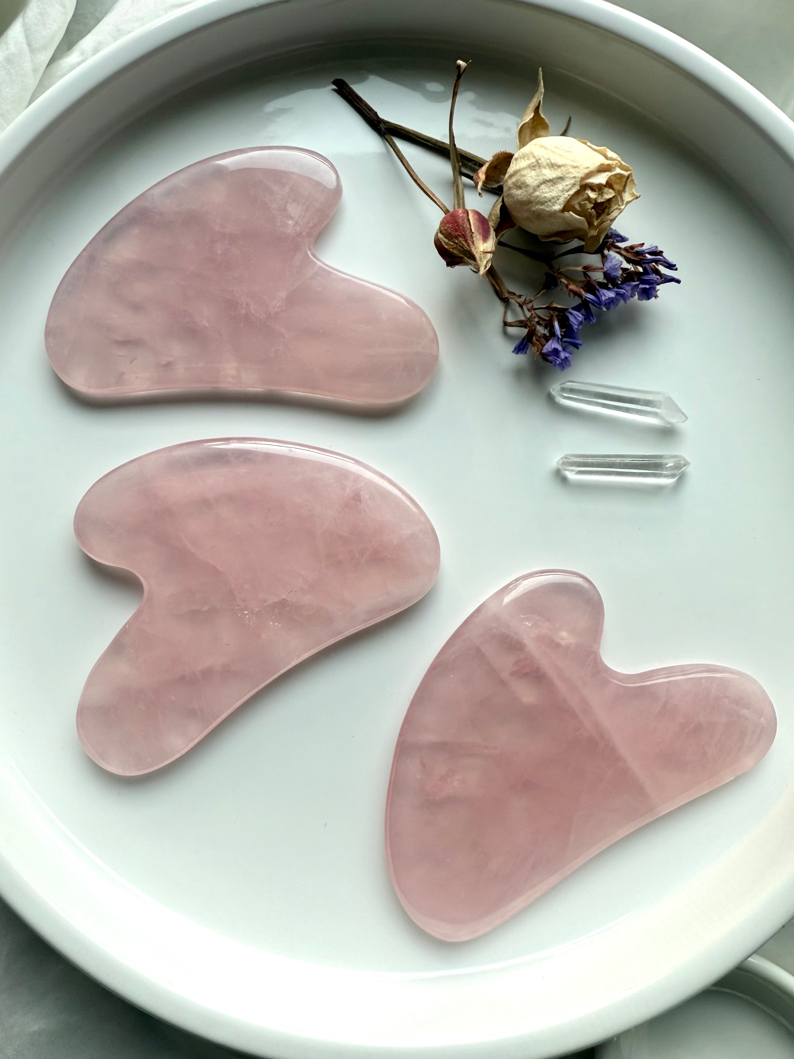 Three Rose Quartz crystal gua sha facial massage tools on white plate with flowers and quartz points