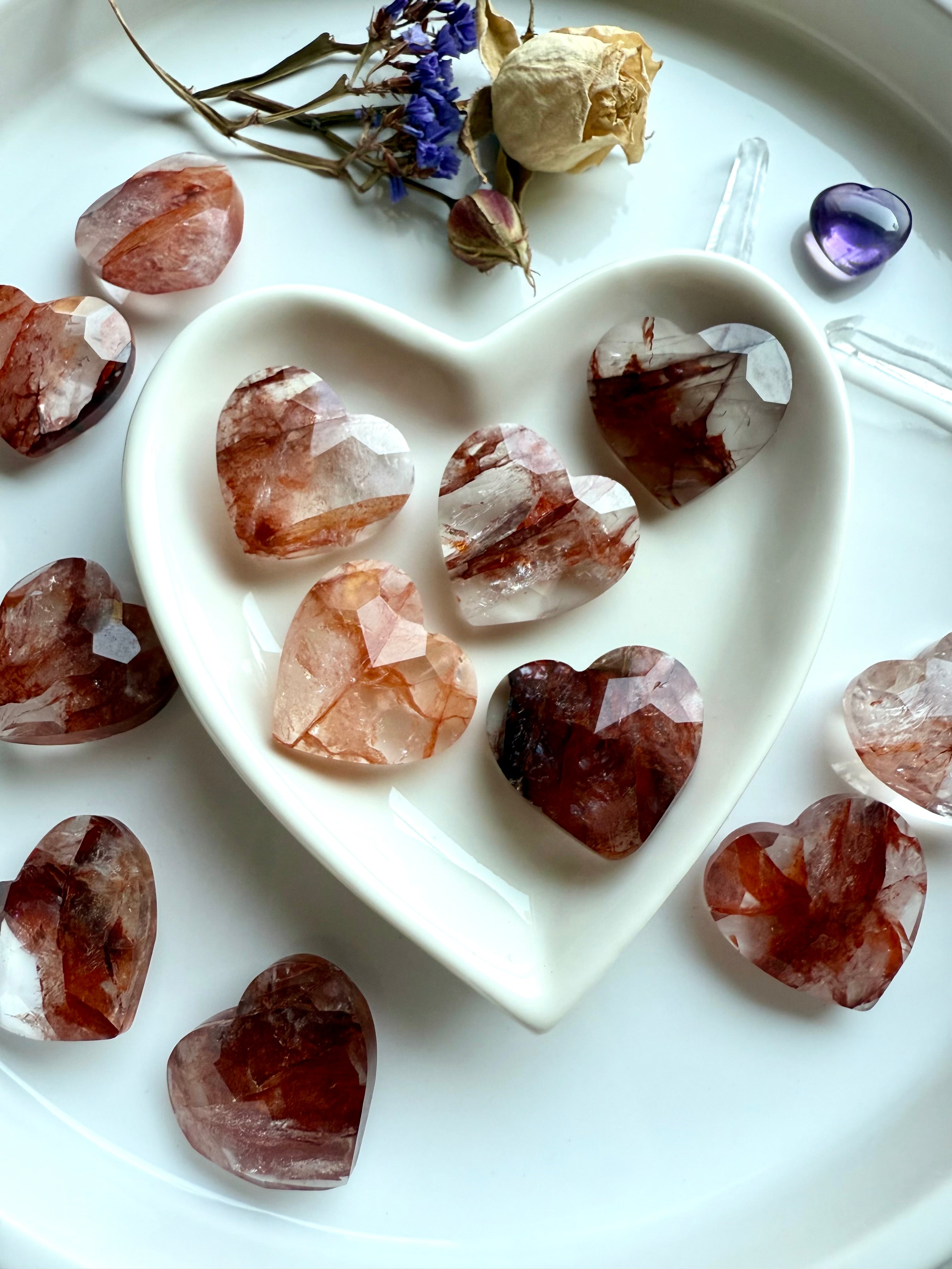Faceted Fire Quartz crystal hearts in a white heart-shaped bowl with flowers, amethyst heart, and clear quartz points – healing crystals for grounding energy