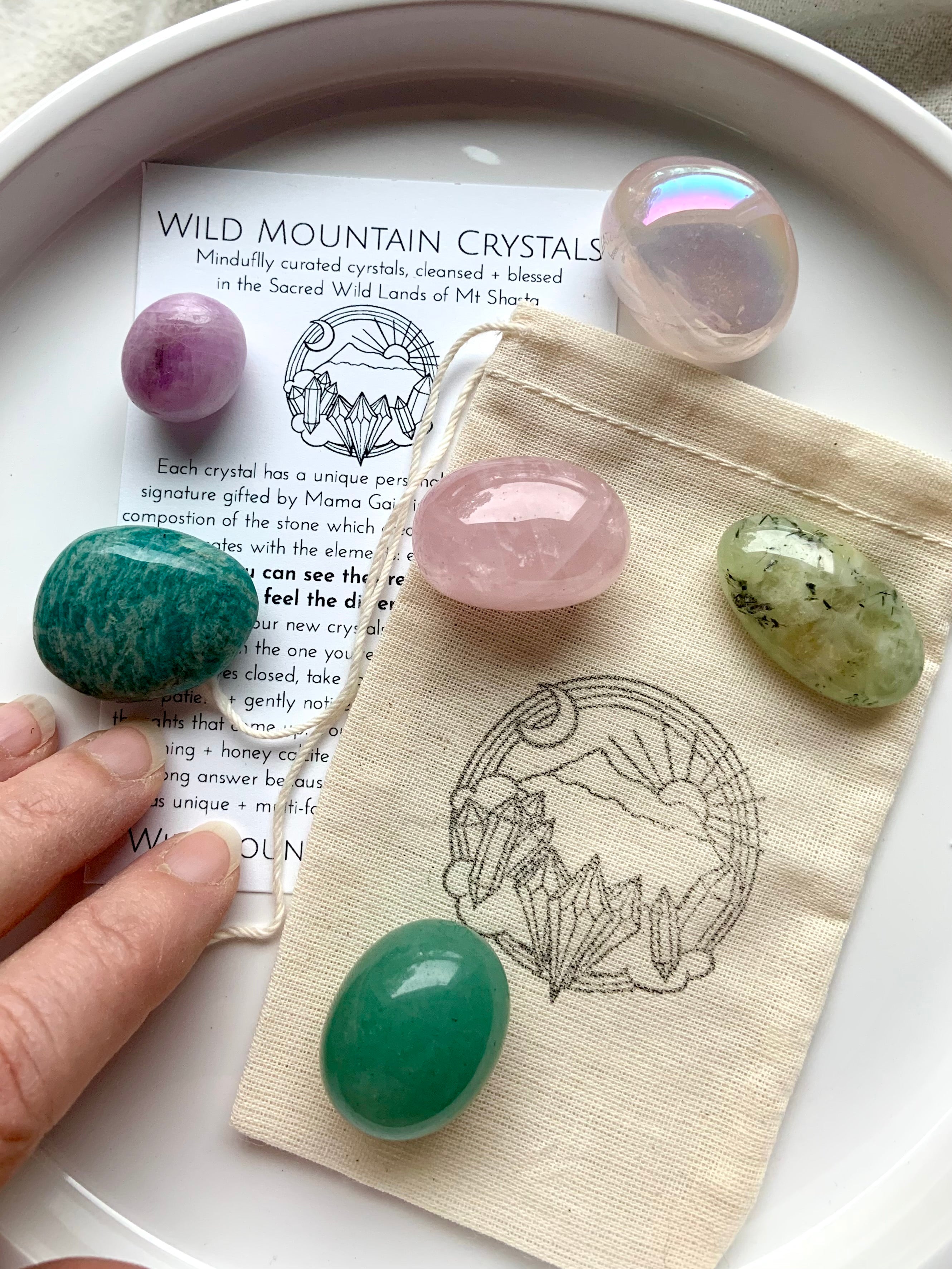 Heart Chakra crystal gift set with 6 pink and green healing stones on top of info card and logo pouch — hand reaching in to select a crystal from the plate.
