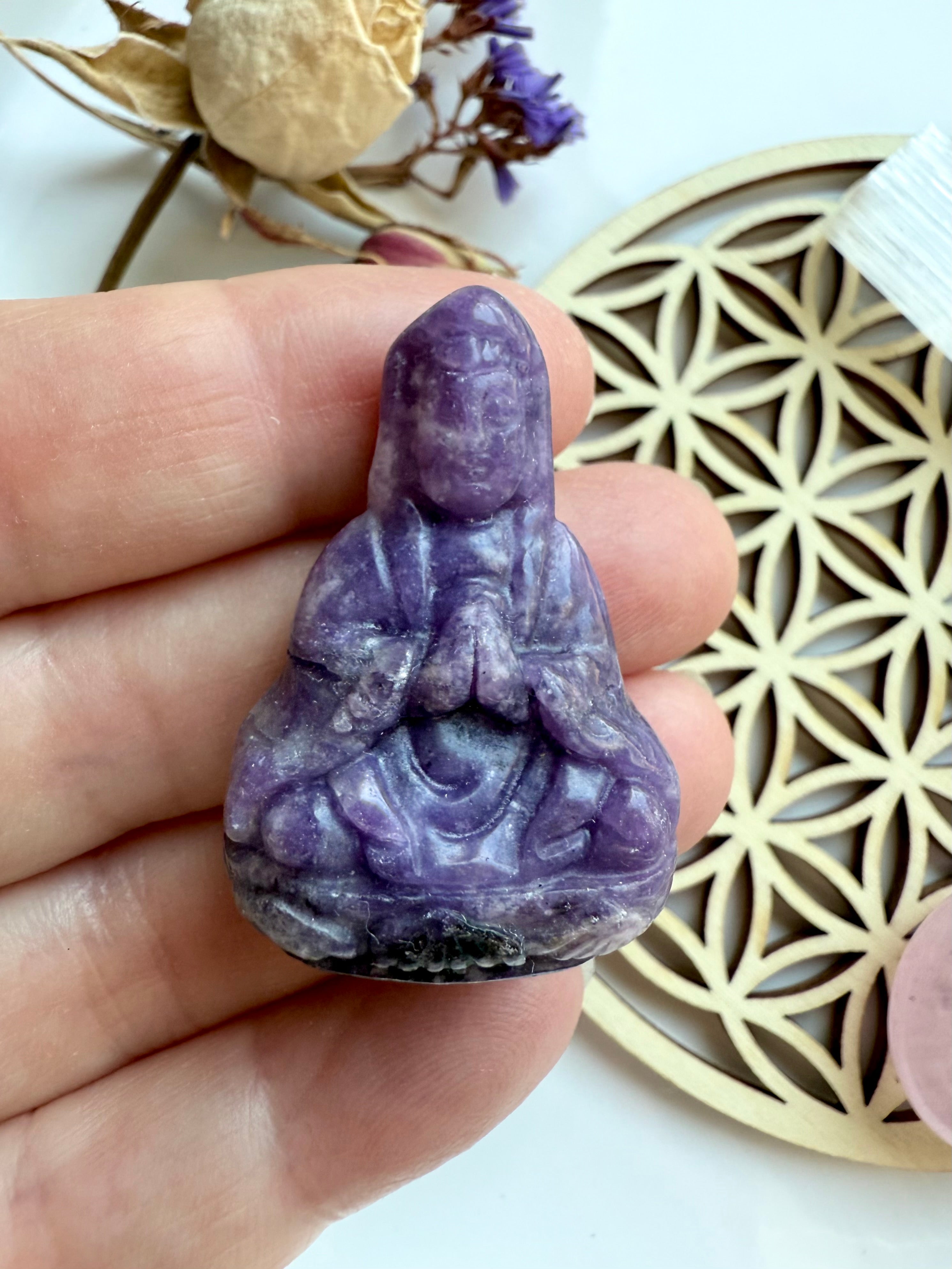 Close-up of lepidolite Quan Yin carving held between fingers with flower of life crystal grid and dried botanicals