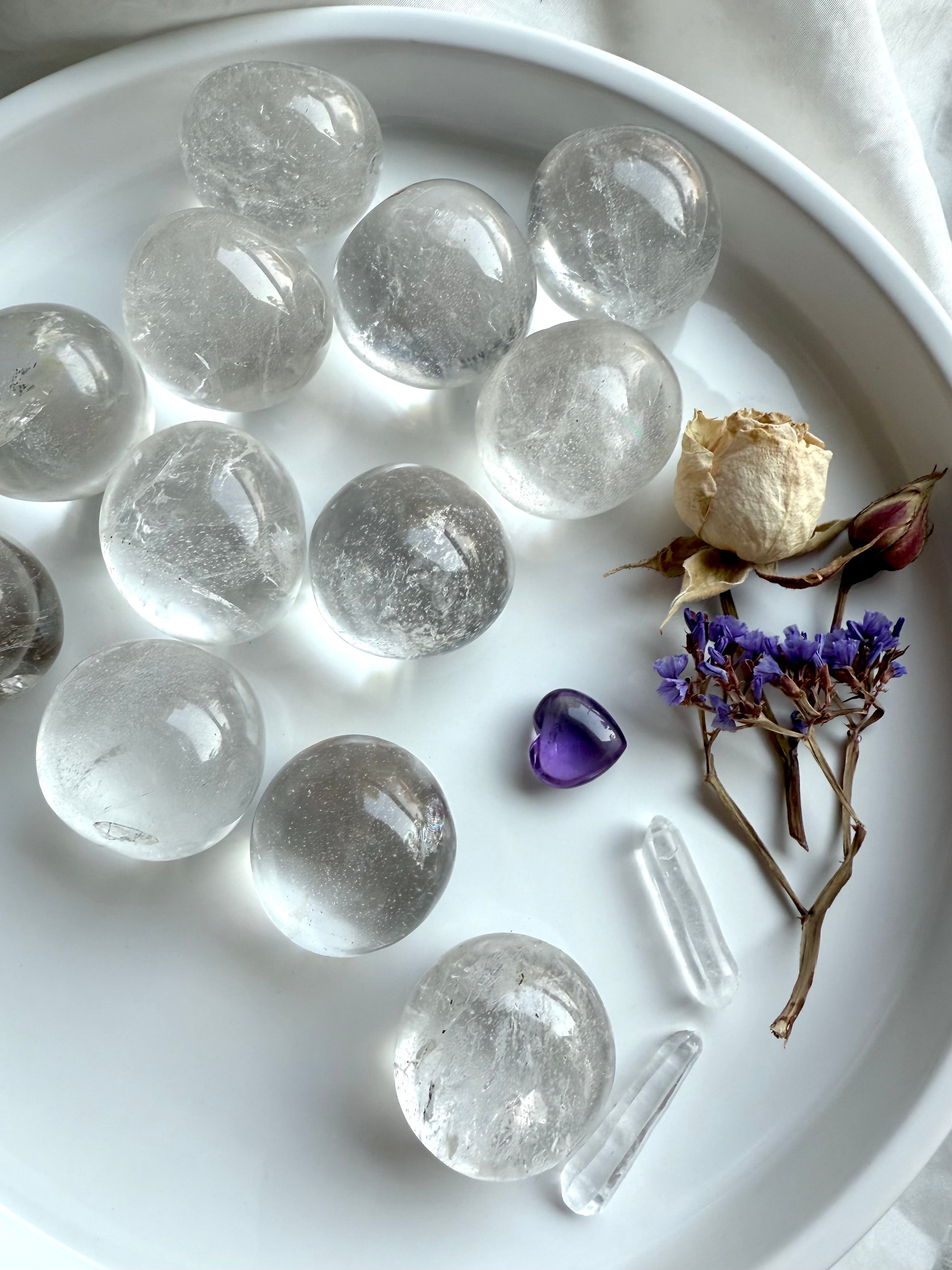 Clear Quartz tumbled stones arranged on a white plate with quartz points, amethyst heart, and dried flower accents