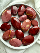 overhead view of polished Mookaite Jasper palm stones on white dish showing earthy red, mauve, cream, and burgundy color variations