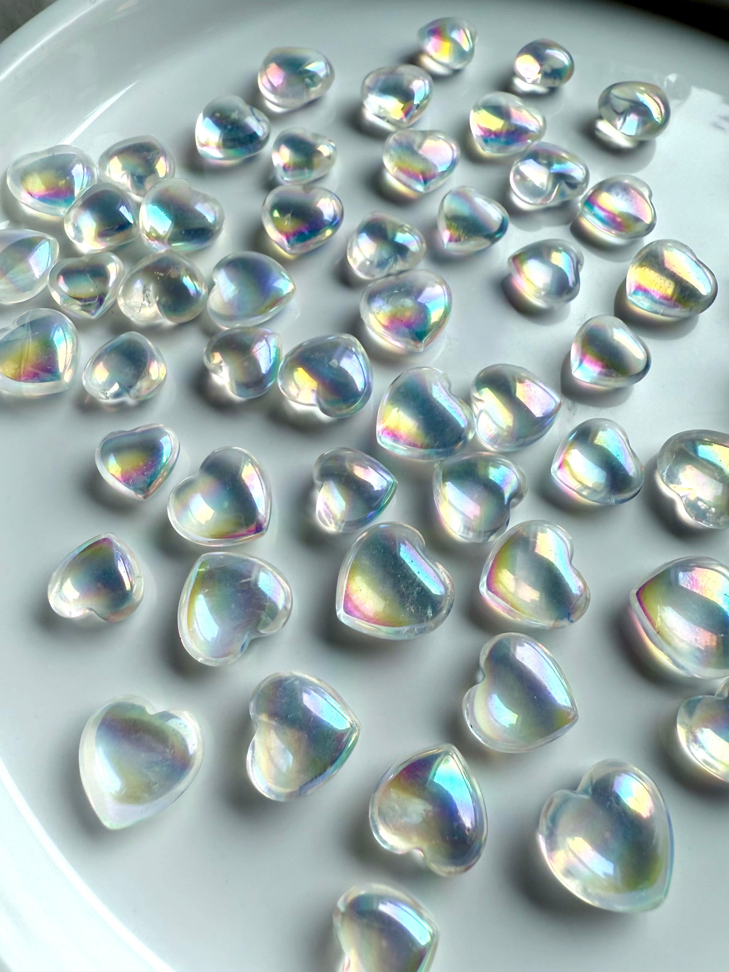Angled view of iridescent mini aura quartz hearts glowing with rainbow light, displayed on white ceramic plate