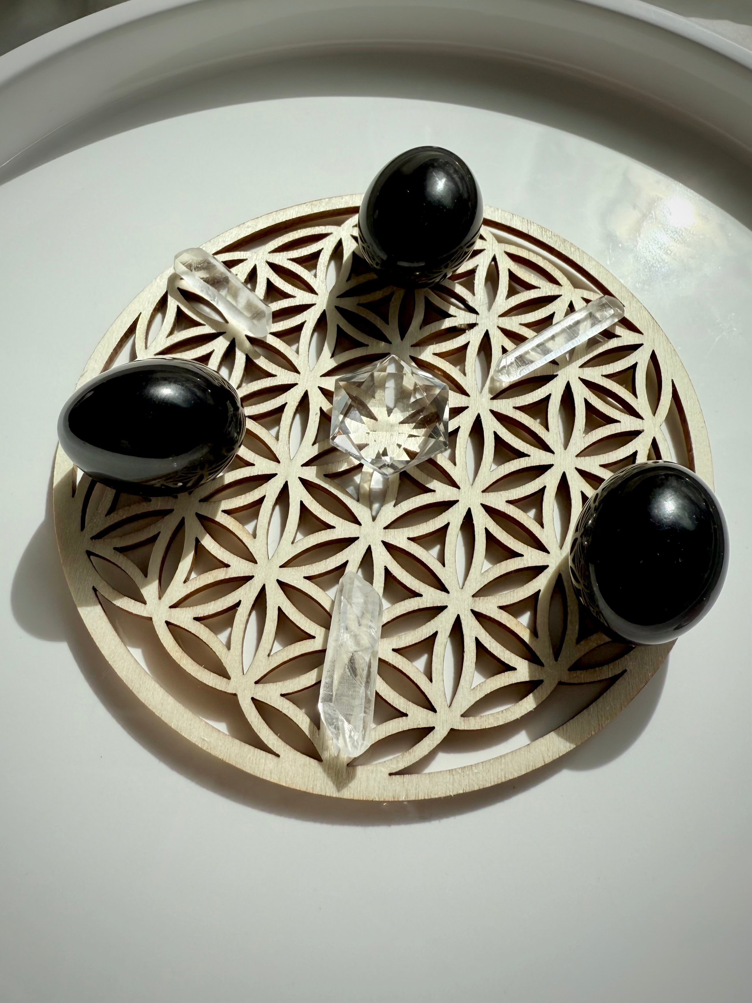 Angled view of crystal grid with small Clear Quartz hexagon centerpiece, arranged with three quartz points and three black obsidian tumbles on a wood Flower of Life grid plate over white surface