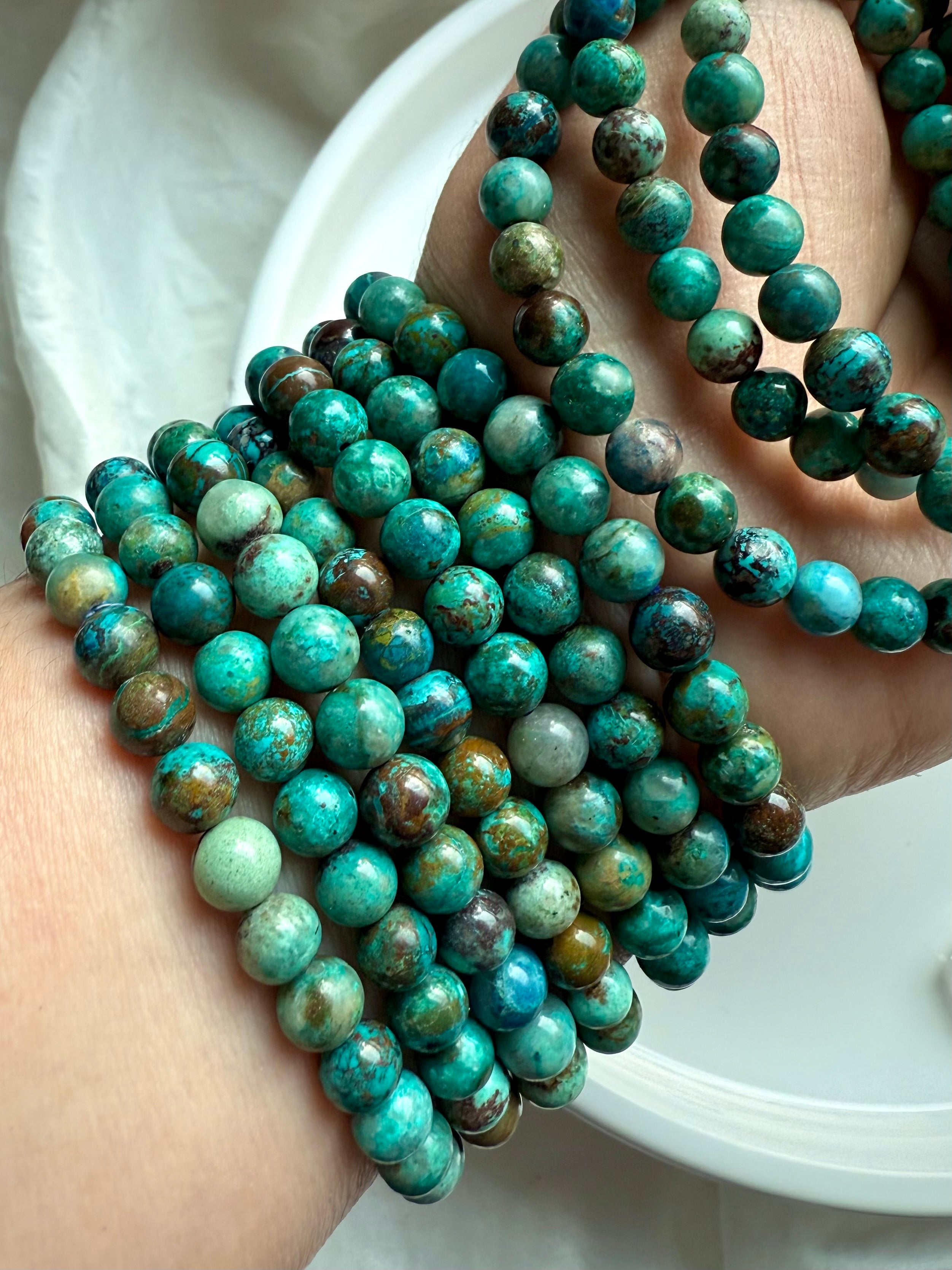 Collection of Chrysocolla Shattuckite stretch bracelets in hand, showing variations of teal, blue, and earthy inclusions - throat chakra healing jewelry