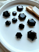 Faceted Black Obsidian hexagon crystals in assorted sizes displayed on a white dish with Palo Santo and Selenite — grounding root chakra stones for protection and energy clearing.