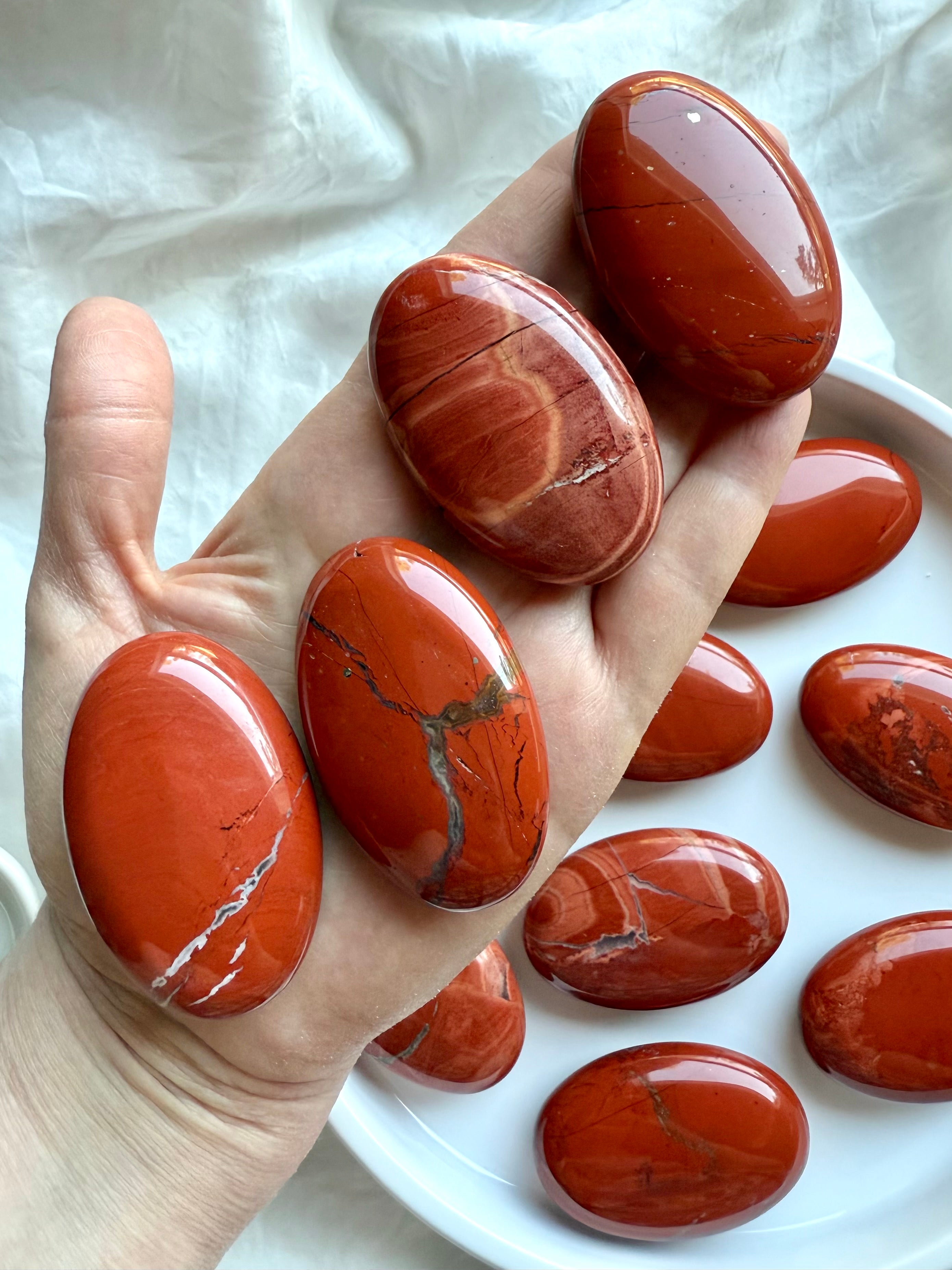 Four Red Jasper palm stones shown on hand above a plate of crystals — earthy healing stones for grounding and strength.