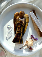 Golden Tiger’s Eye angel carving displayed in a white dish with Selenite, Palo Santo, and dried flowers