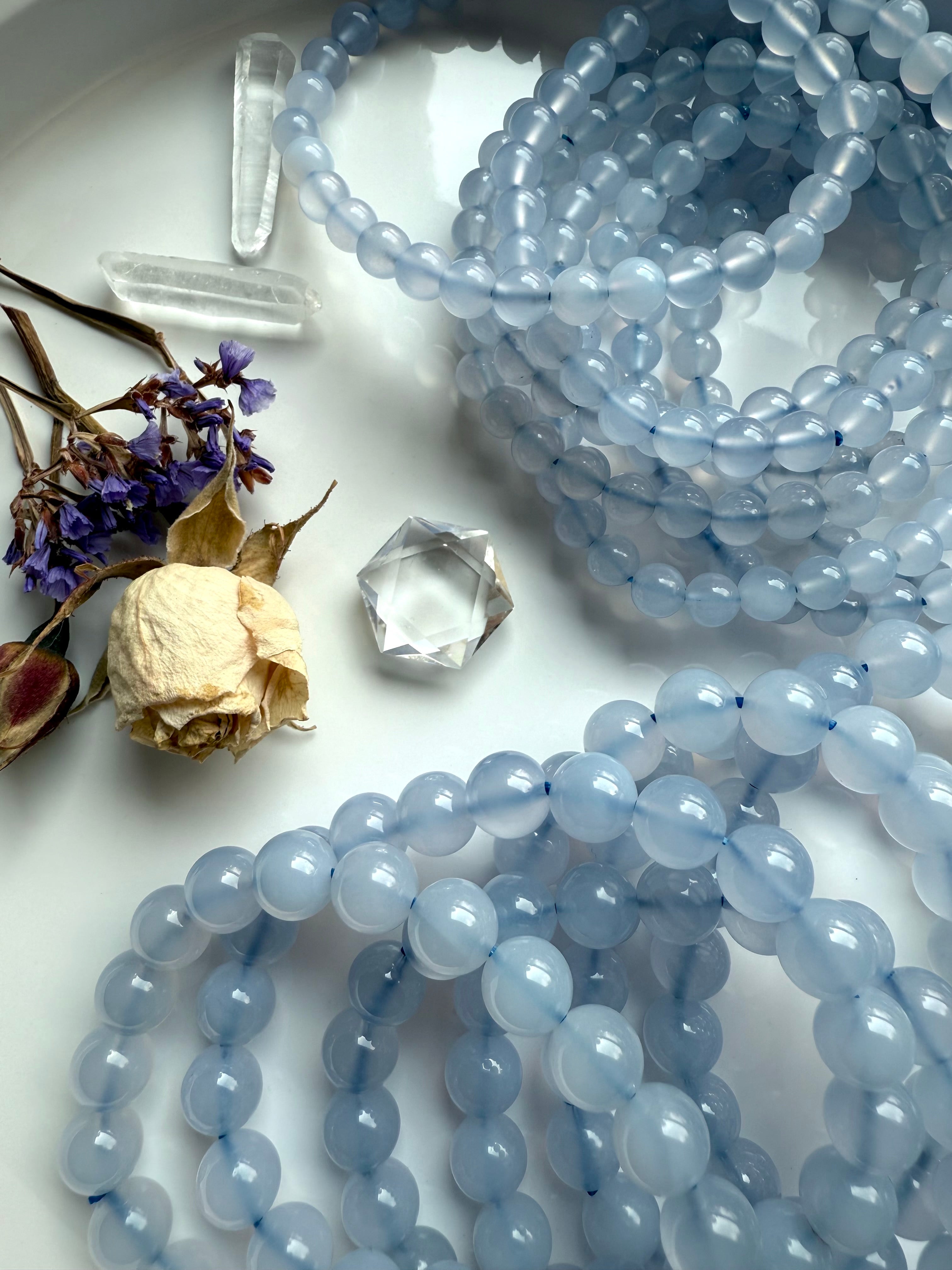 Blue Chalcedony bead bracelets displayed in gentle coils on a white ceramic plate with dried flowers — soothing crystal jewelry for calm and clarity.