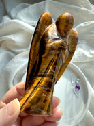 Side view of Tiger’s Eye crystal angel carving glowing in natural light, showcasing chatoyant bands and protective wings