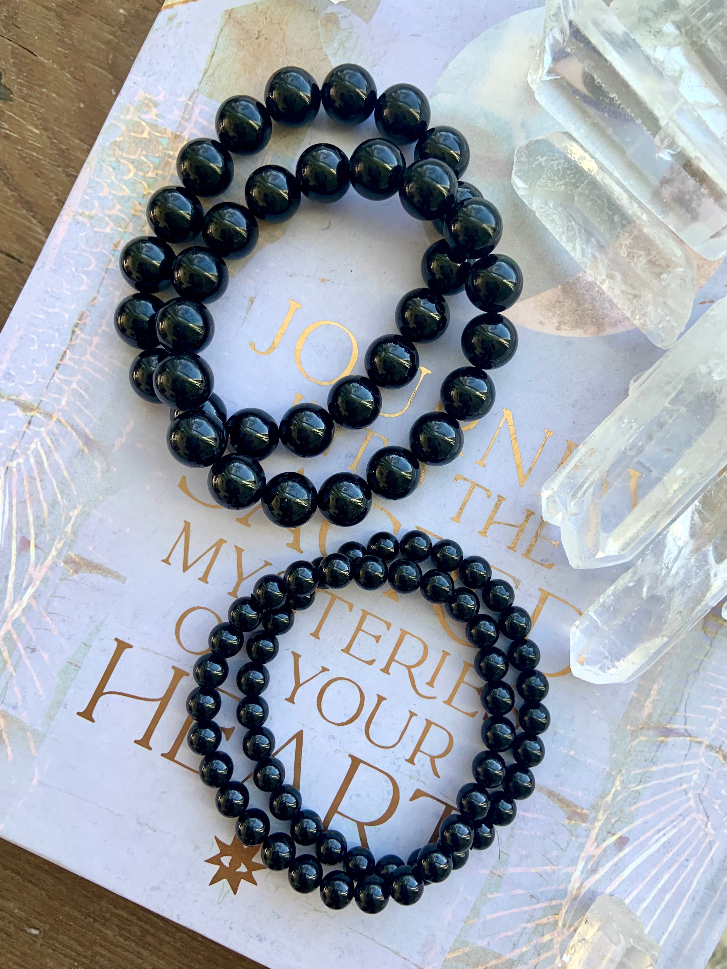 Two sizes of Black Tourmaline bracelets next to clear quartz points in a flatlay — glossy 10–11mm and 6–7mm bead sizes shown for comparison