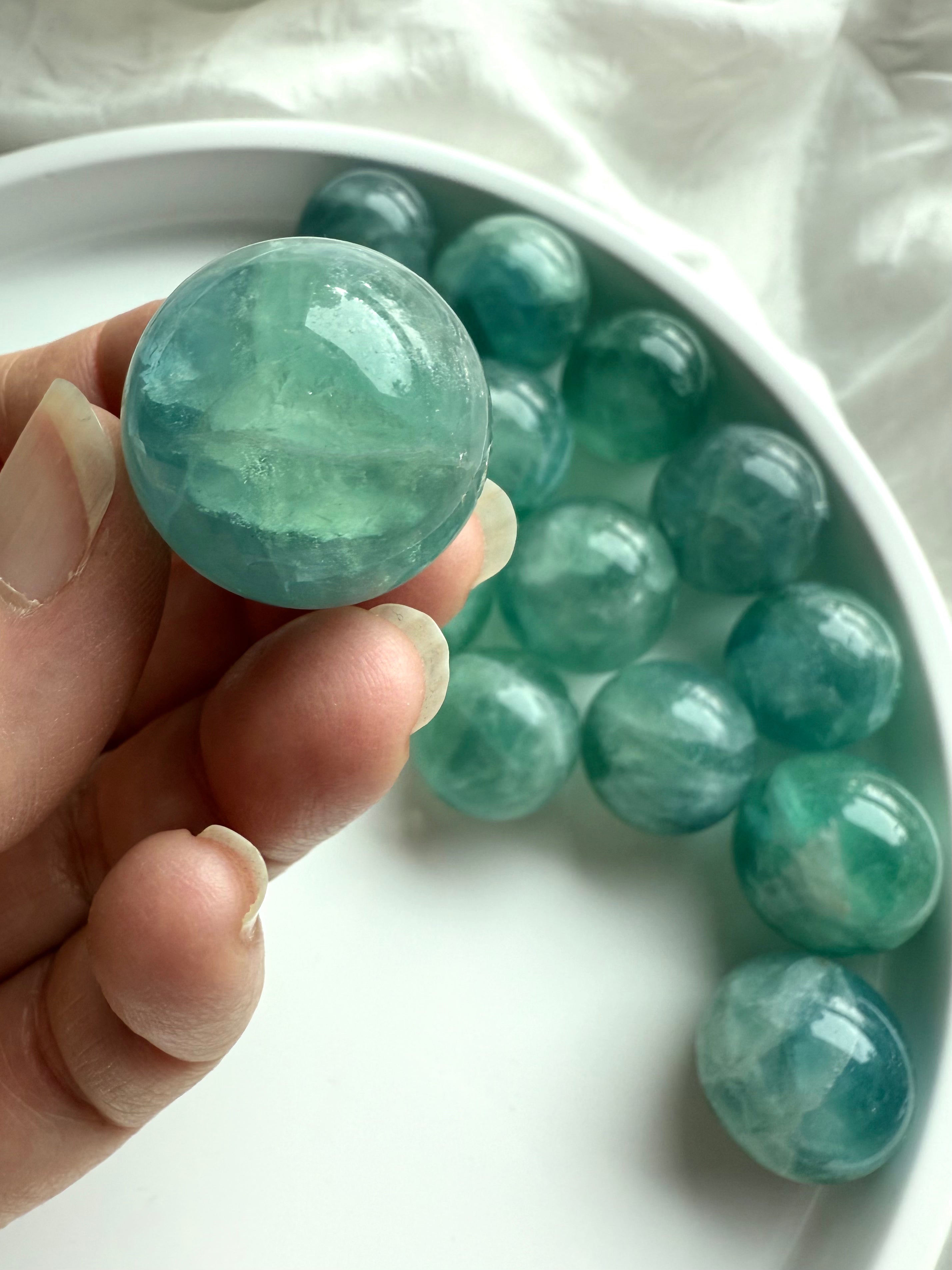 Hand holding a polished teal Fluorite crystal ball showing translucent blue-green patterns for meditation and chakra healing.