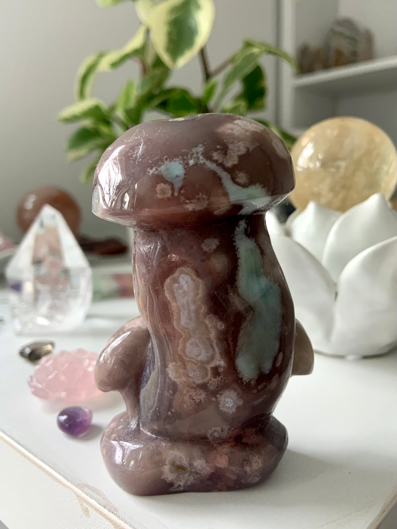 Black Flower Agate Mushroom Carving shown within a crystal collection and a plant behind it