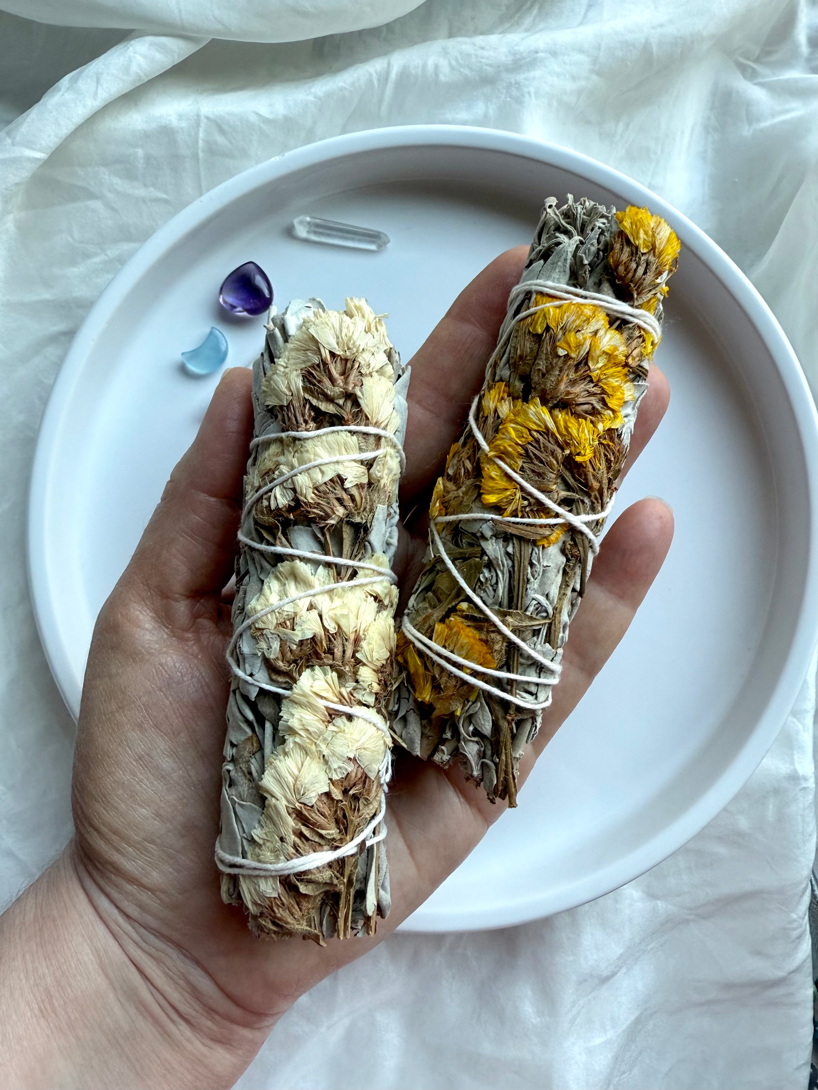 Hand holding two white sage bundles wrapped with yellow and white statice flowers for energy cleansing and spiritual smudging 